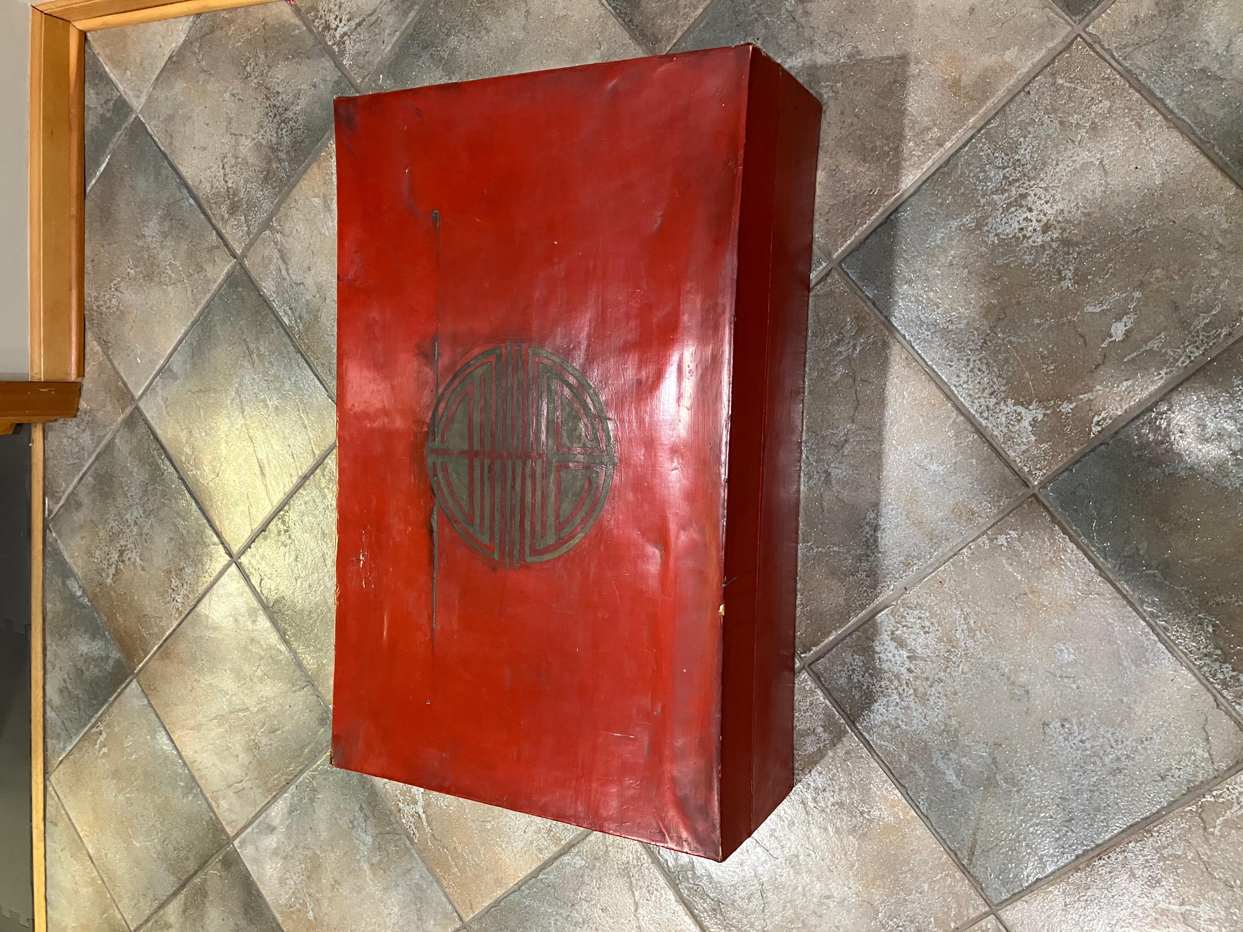A richly colored red lacquered Chinese storage trunk, dating to the early 20th century, featuring original brass hardware and hand-painted naturalistic decorations. The lid showcases a large circular seal medallion, while the side panels are adorned