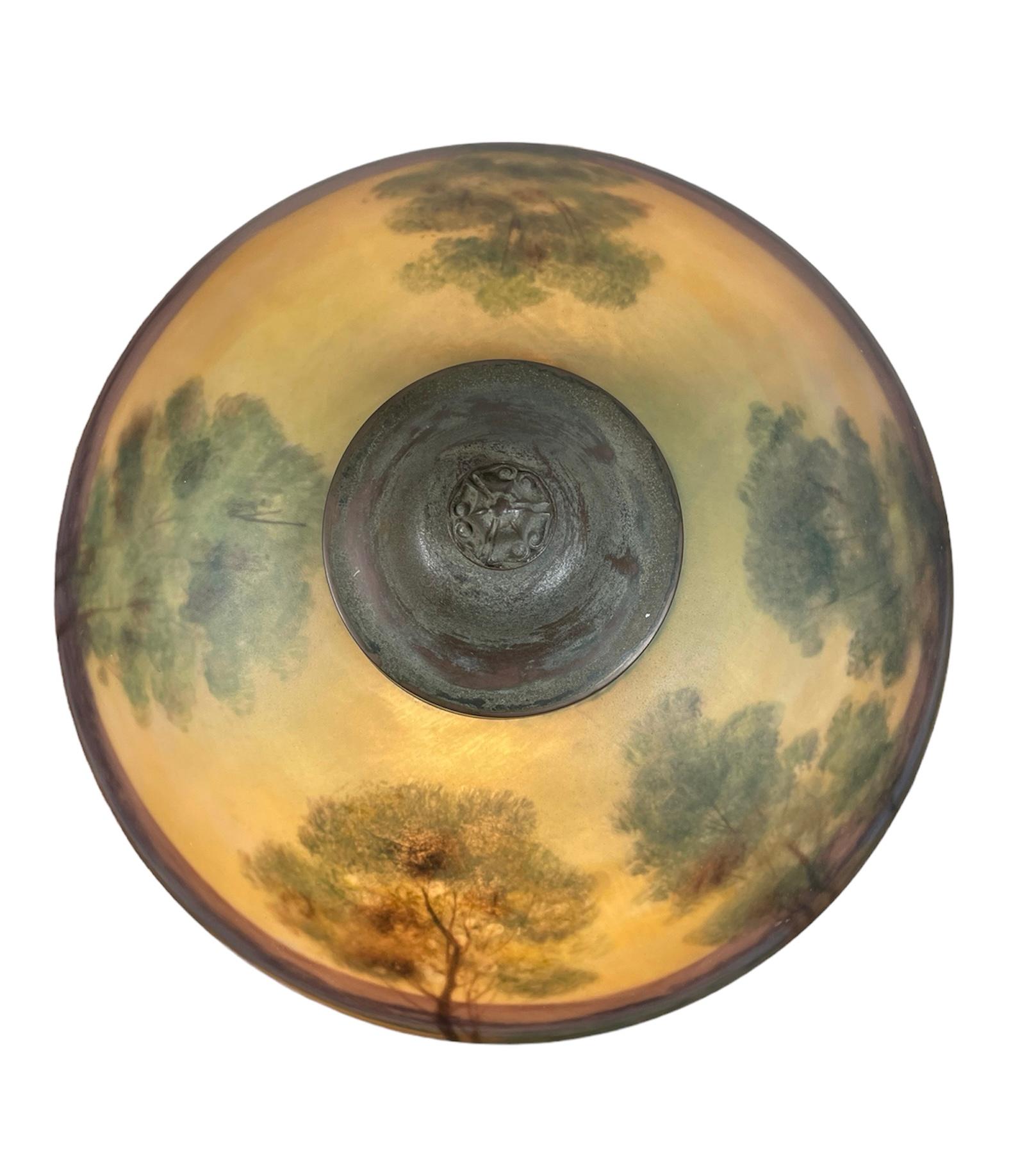 This is a Reverse Painting Dome Glass Shade and Bronze Table Lamp. It depicts a landscape with several tall green trees, dry grass and a lake. The background has blue, brown, green and purple color shades. The heavy iron metal stand of the lamp is