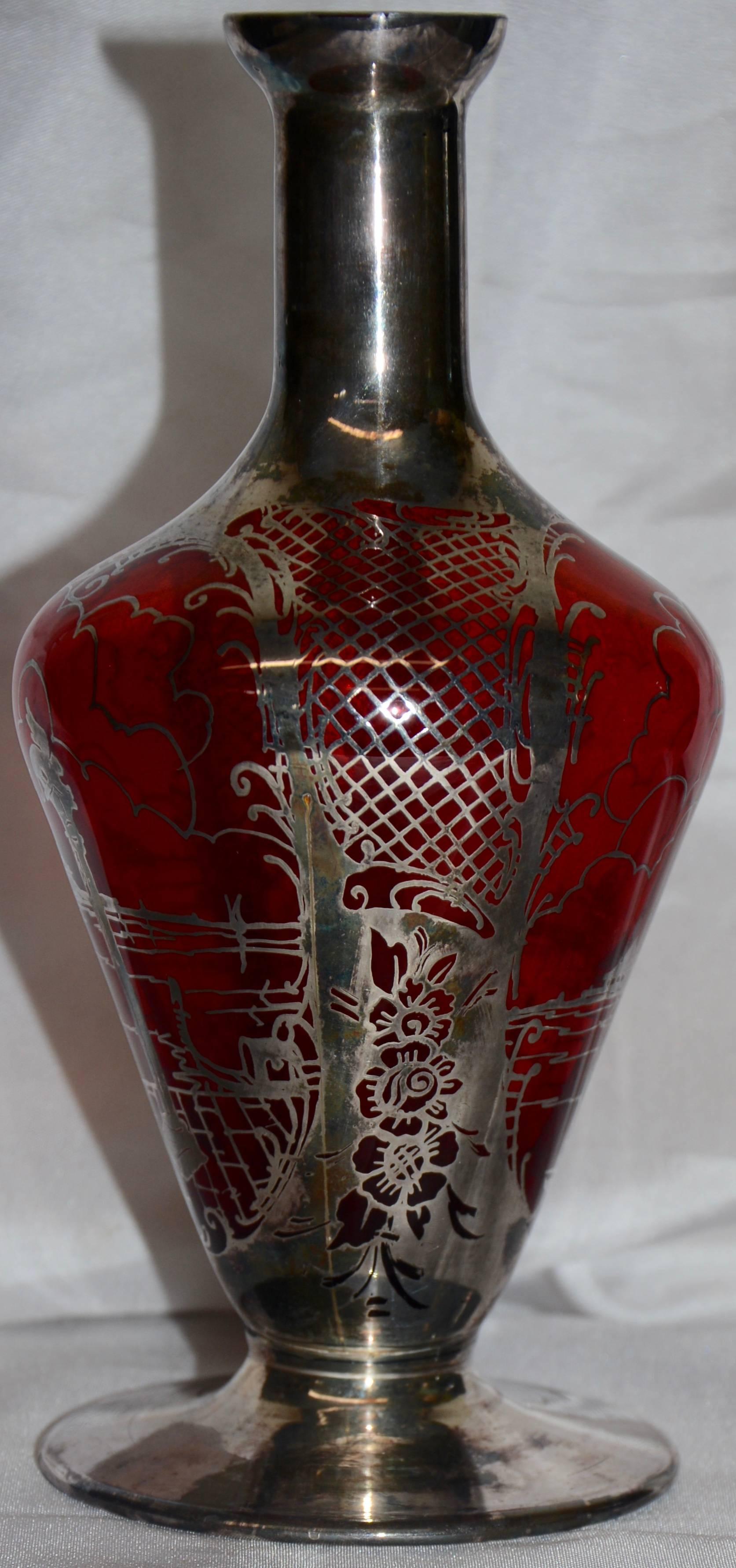 This is a lovely Art Nouveau vase formed of ruby red glass with a delicate silver overlay. Dainty lattice work tops an array of flowers on two sides and an outdoor scene is captured on the other two sides.