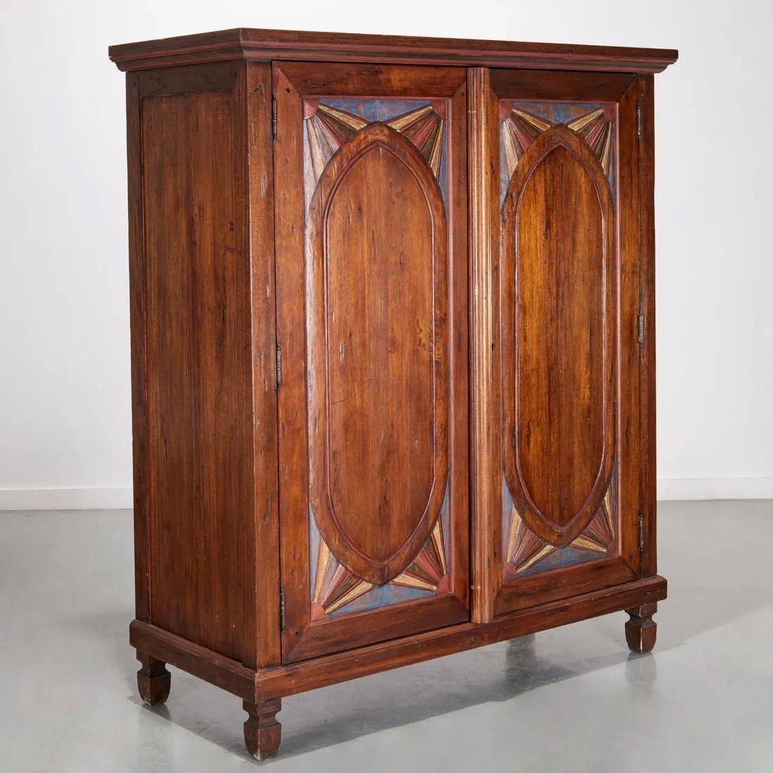 Un armadio in stile folk del XX secolo, affascinante e ricco di charme, con decorazioni policrome dipinte a mano. Realizzato in legno massiccio, questo mobile presenta splendidi pannelli ad arco sulle doppie ante con vivaci motivi a raggiera nei