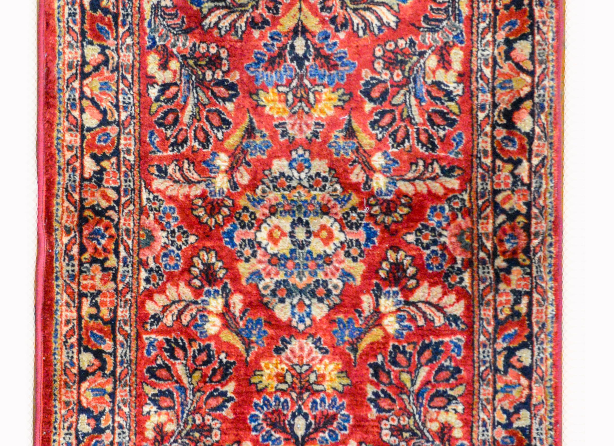 A gorgeous early 20th century Persian Sarouk rug with an all-over pattern containing several large flower clusters woven in indigo, pink, gold, and white, against a dark cranberry background. The border is wonderful with a central floral patterned