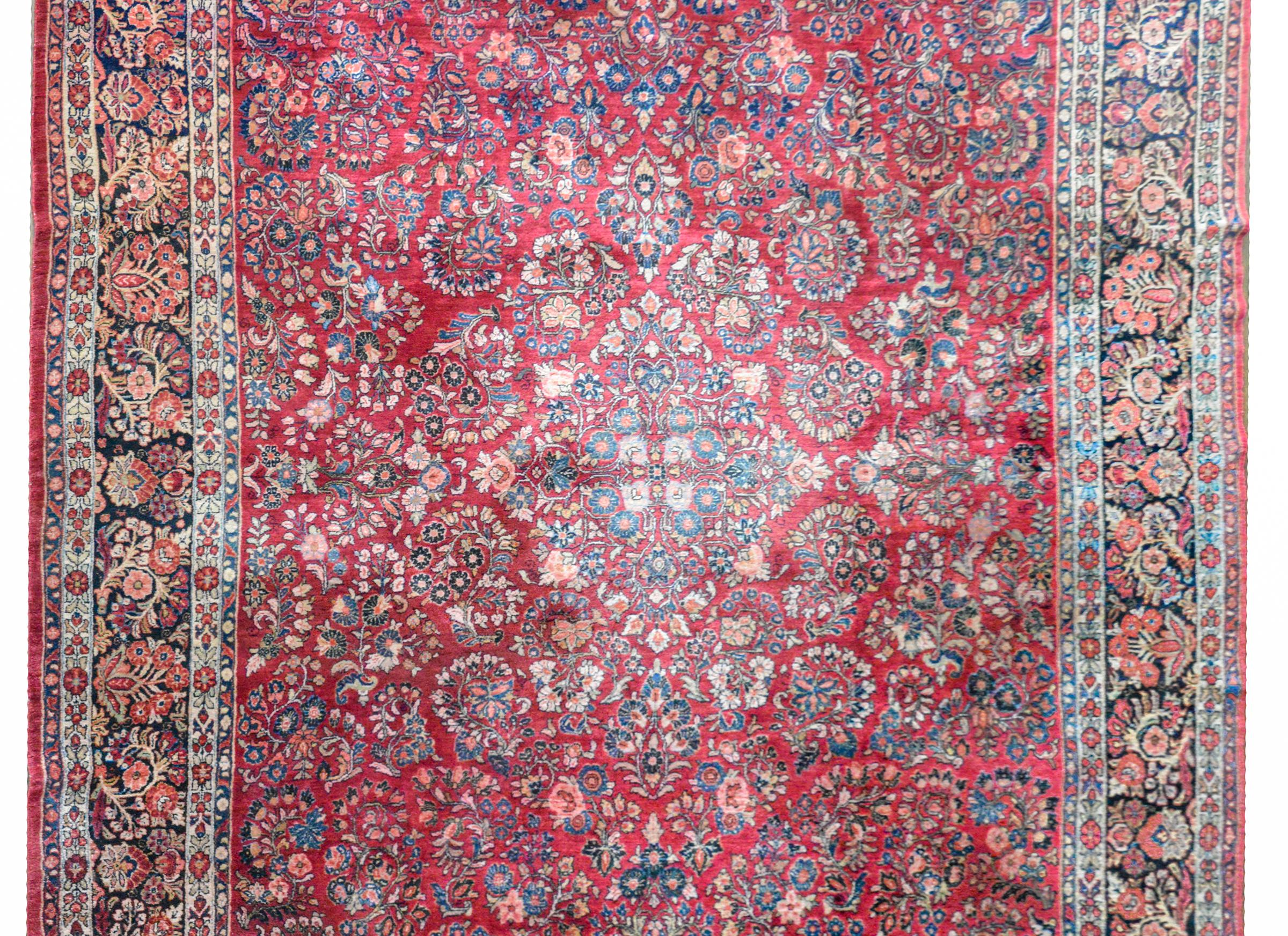 A early 20th century Persian Sarouk rug with a traditional pattern containing myriad clusters of flowers woven in light and dark indigo, cream, pink, and green, set against a crimson background. The border is wide, with a central floral patterned