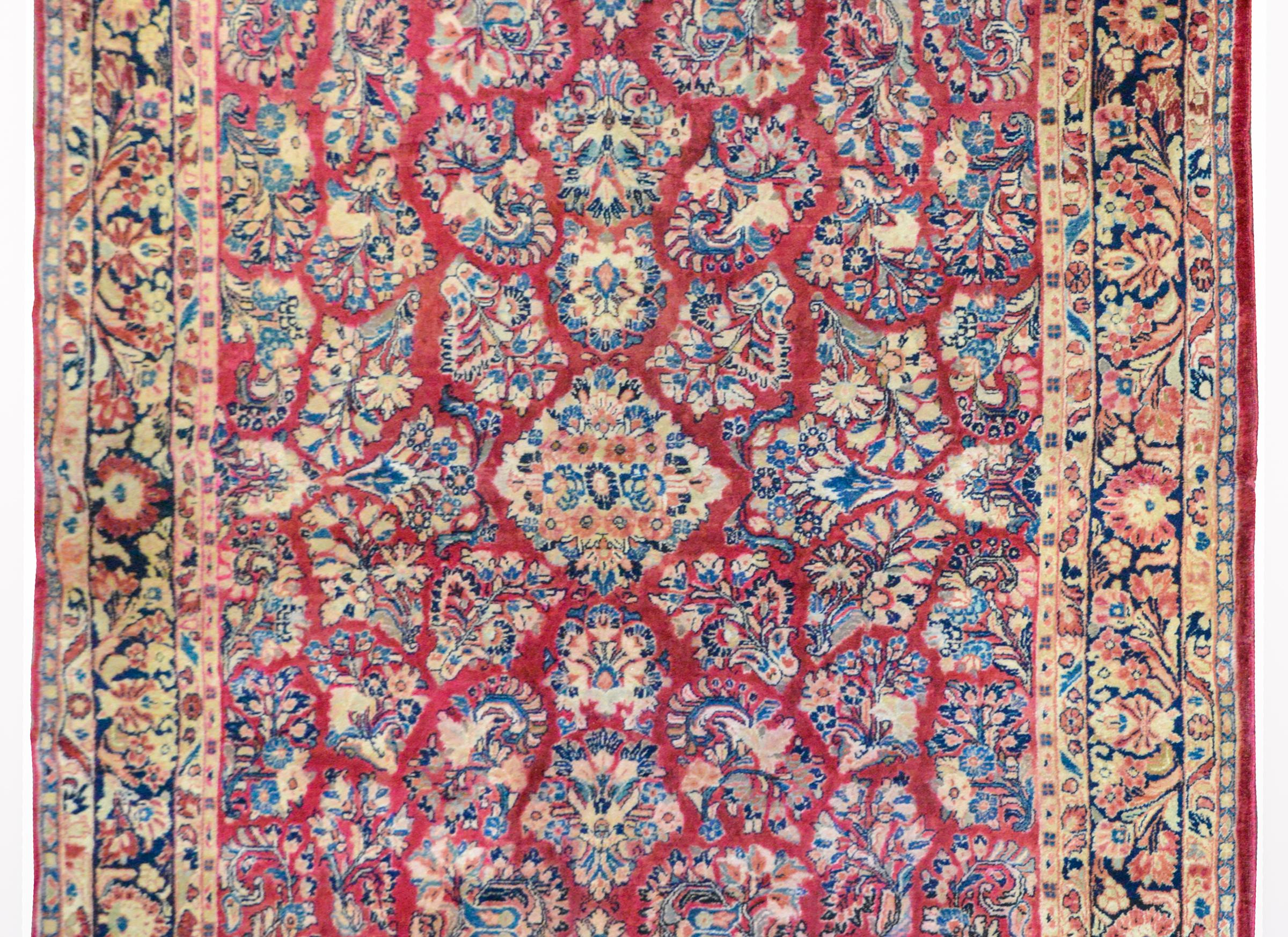 A beautiful early 20th century Persian Sarouk rug with an all-over mirrored floral cluster pattern woven in light and dark indigo, cream, and orange, against a cranberry field and surrounded by a large-scale floral partnered border, flanked by