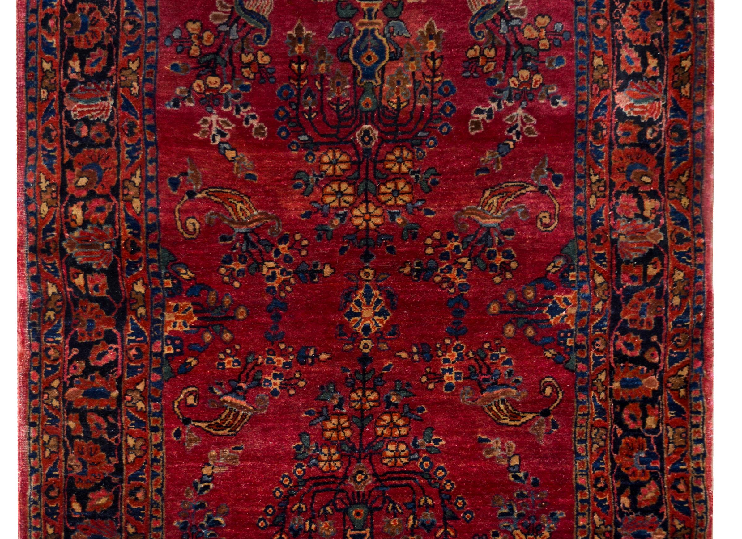 A beautiful early 20th century Persian Sarouk Mohajeran rug with a mirrored tree-of-life pattern woven in indigo, cream, and pink, against a dark cranberry background, and surrounded by a wide floral and scrolling vine patterned border.