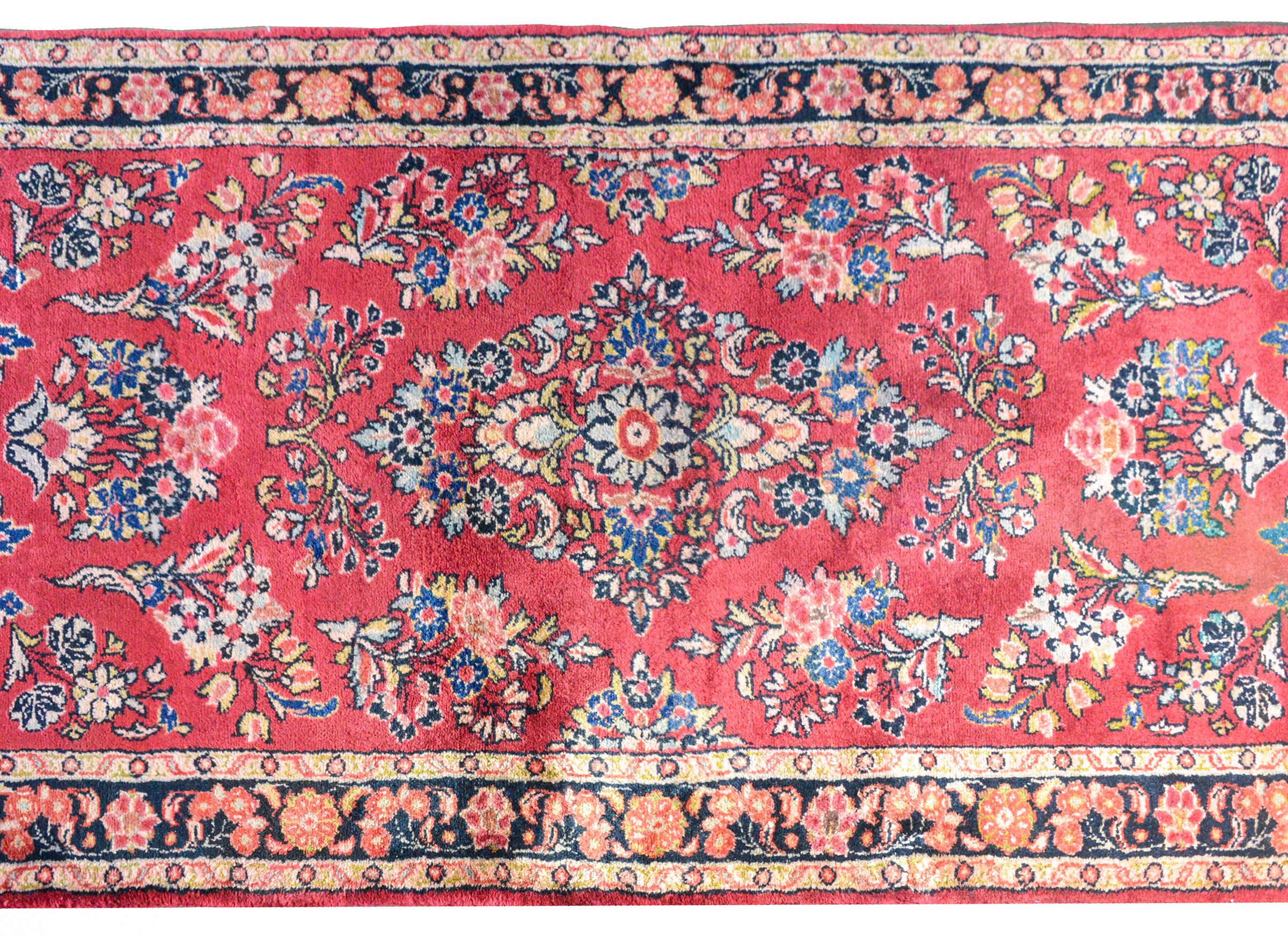 A wonderful early 20th century Persian Sarouk runner with a beautiful mirrored floral pattern woven in pink, gold, light and dark indigo, and white, against a bright red background. The border is simple with several petite floral patterned stripes.