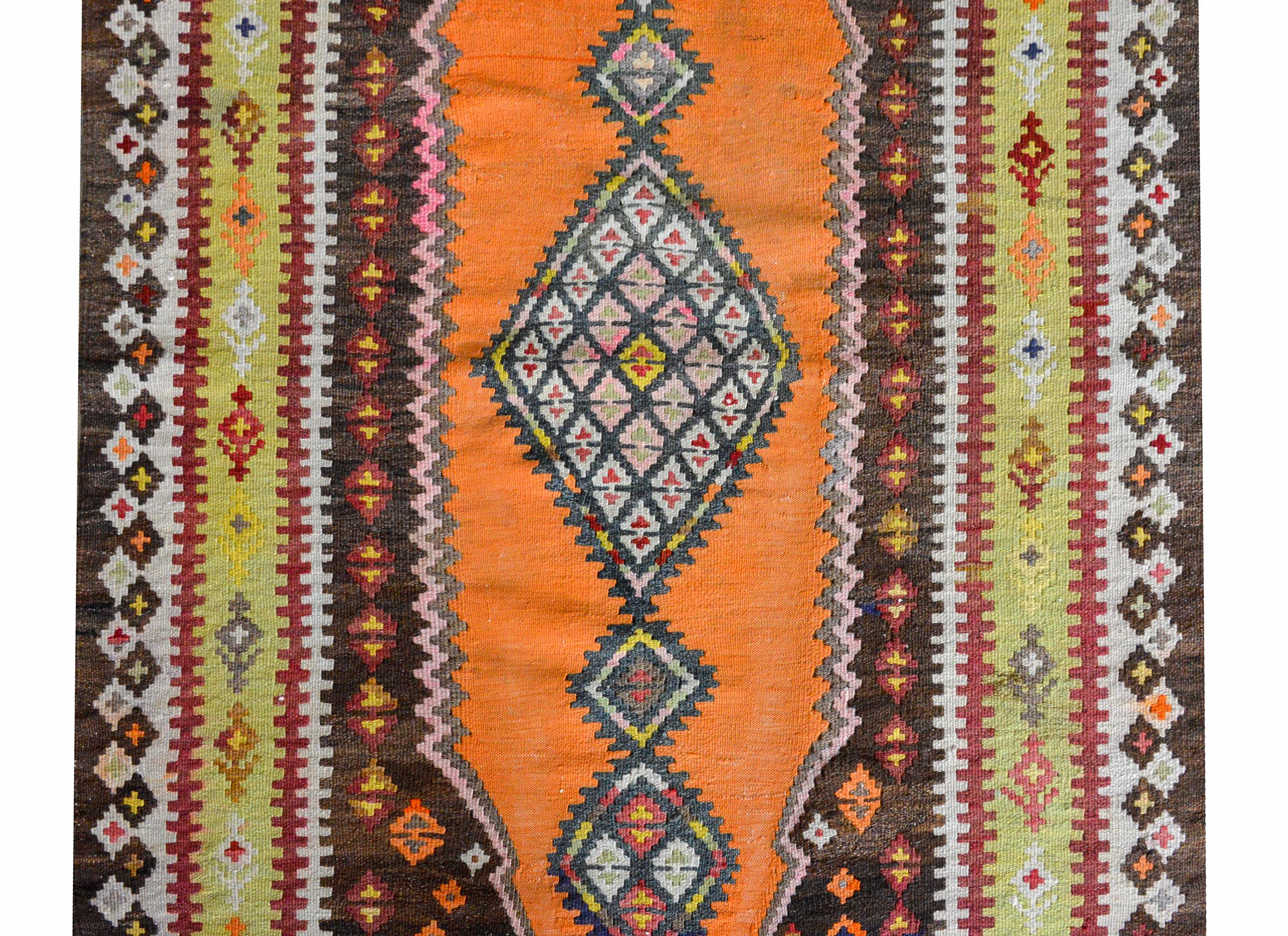 A wonderful early 20th century Persian Saveh Kilim rug with a tribal pattern containing multiple diamond motifs set against an orange background, and surrounded by a wide border composed with multiple floral and geometric pattered stripes.