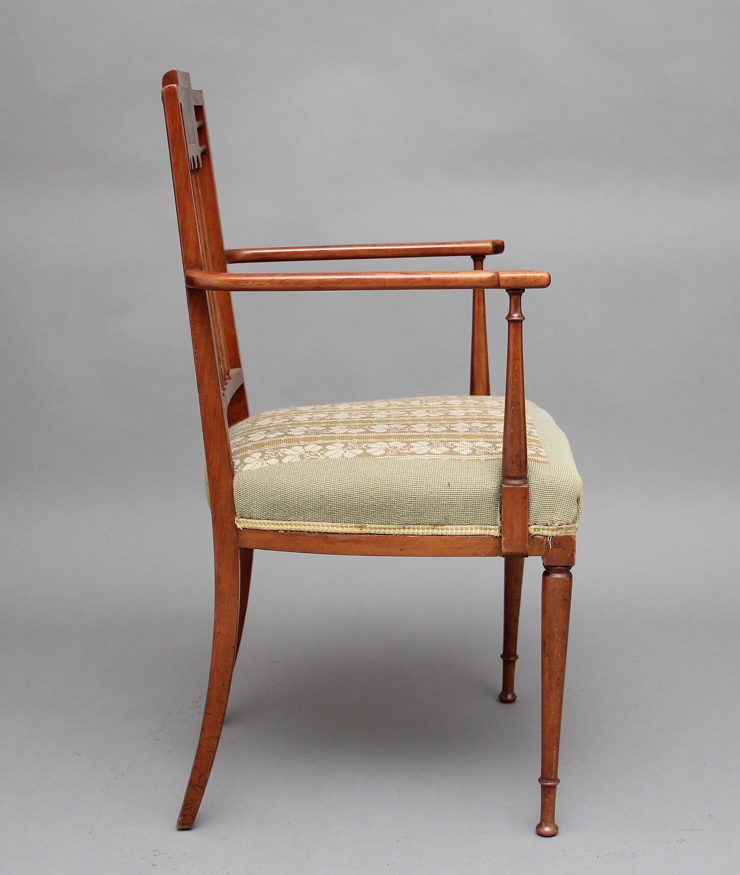 An early 20th century Sheraton revival satinwood armchair painted overall with floral decoration, the chair back consisting of finely turned spindles with a panel at the top painted with cherubs, turned uprights with shaped arm supports, embroidered