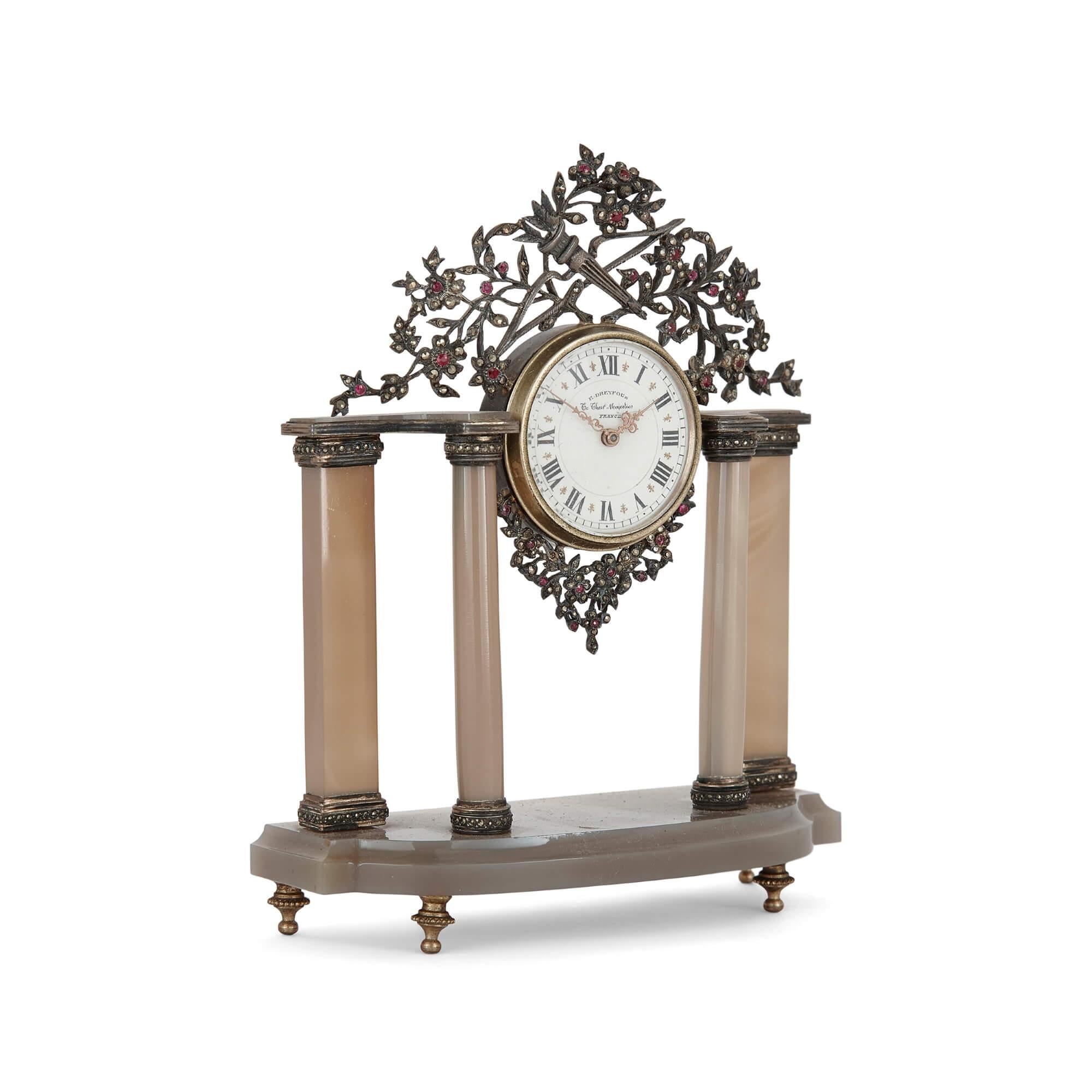 This delicate table clock features a surmounting design of silver foliate and flowers, which are mounted with precious stones. Held centrally within the silver flowers is the circular clock dial, which features a gilt border and black Roman numerals