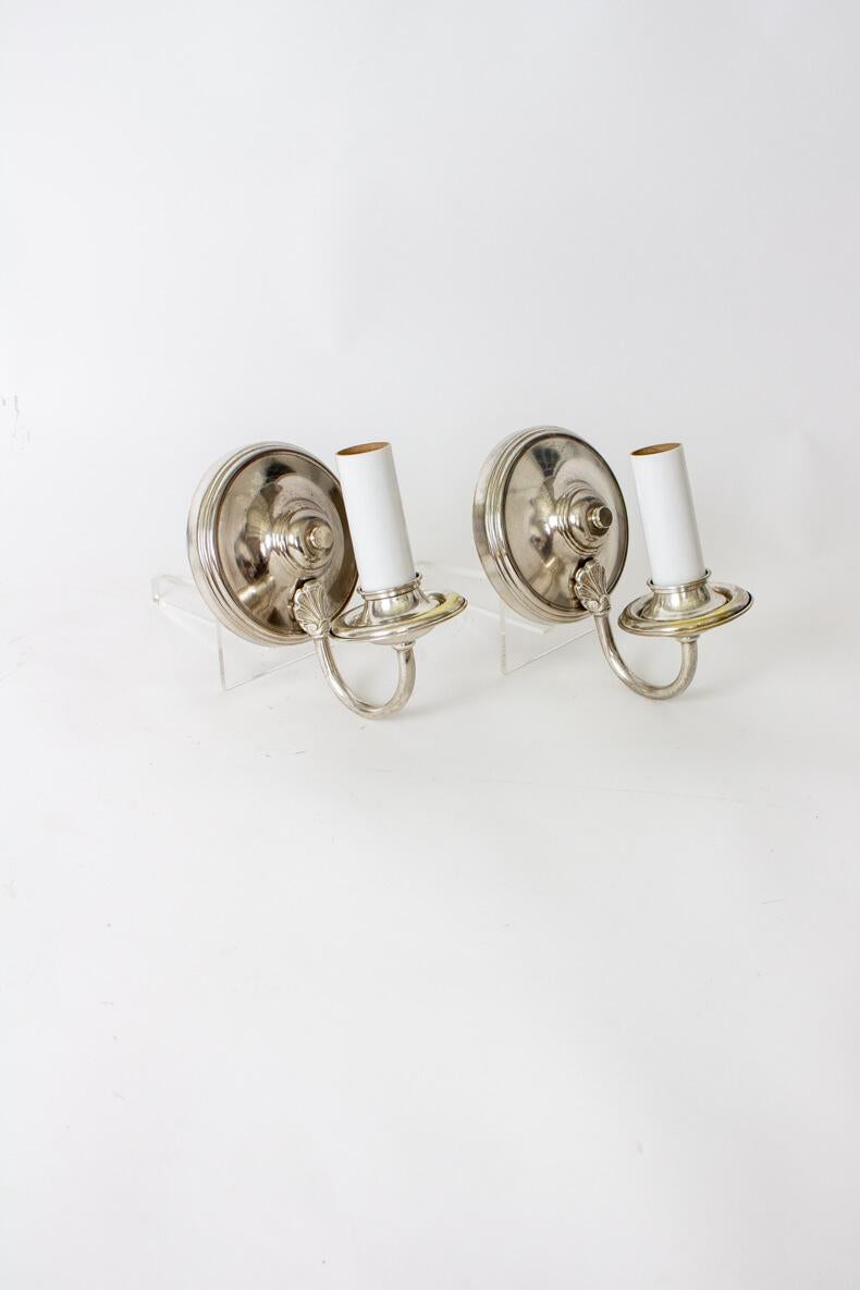 A pair of elegant and simple silver sconces. Round backplate with a single curved arm and white candle covers. Takes a standard Edison base socket, and can be used with or without a shade. Backplate is 5” diameter. Silver has been polished and is in