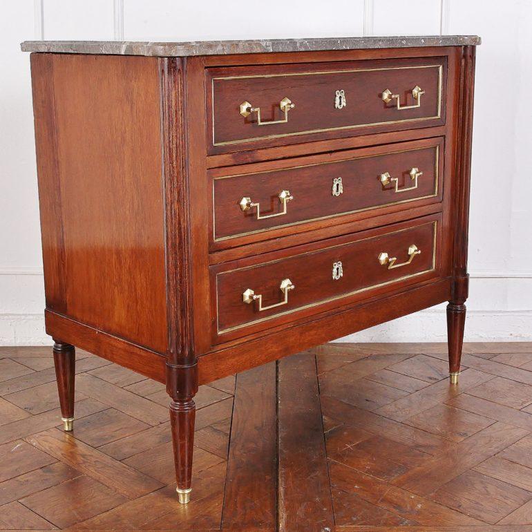 A smaller-scaled early 20th century directiore style commode in mahogany with fluted column accents at each front corner and an original marble top. The three drawers have polished brass trim and key plates, the turned tapering legs are also