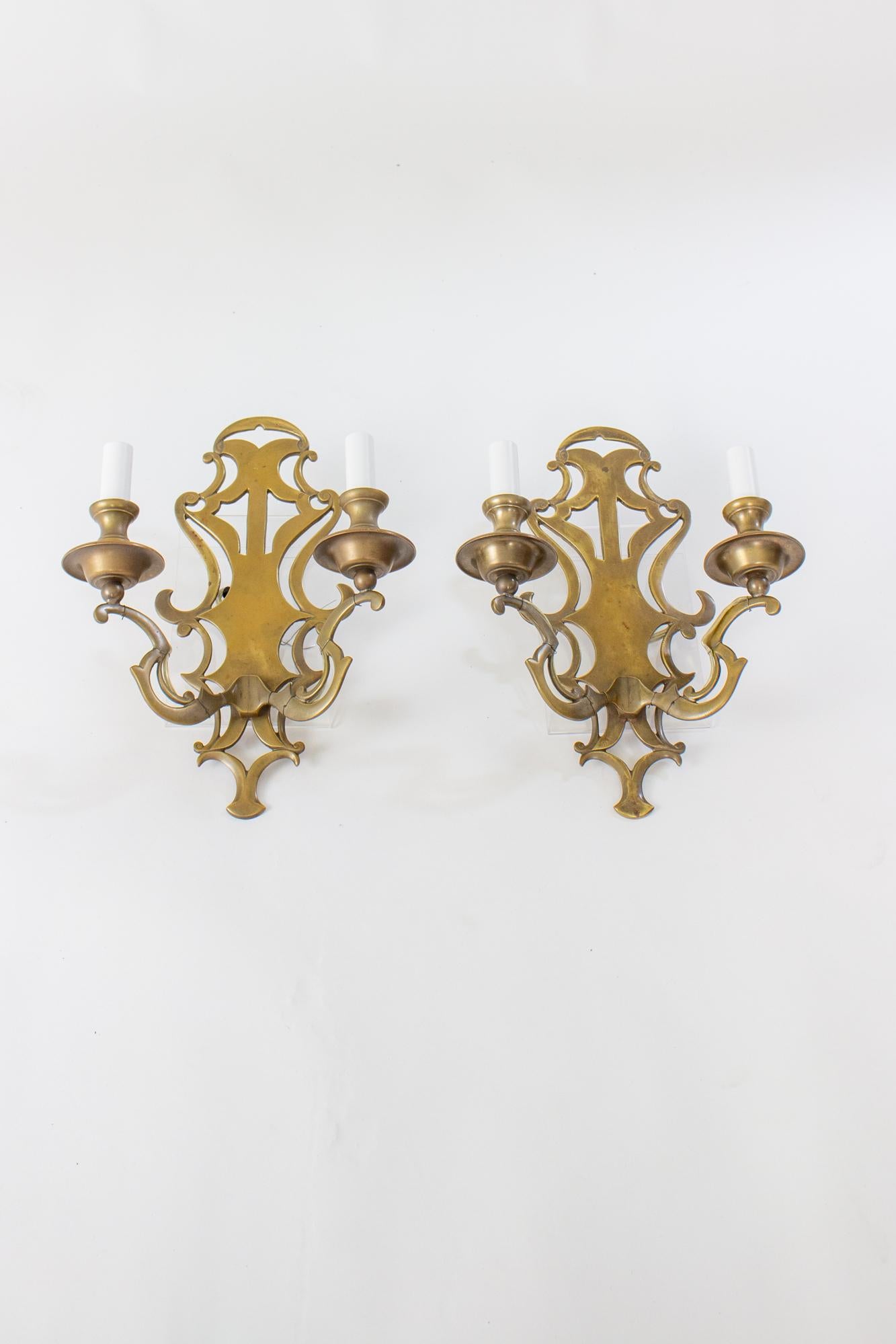 Early 20th Century solid brass sconces, a pair. Flat backplates with cut out pattern. Two arms. All in solid brass with an even natural patina. Rewired with new candlecovers. Originally designed to hang from wall from the top curved element on the