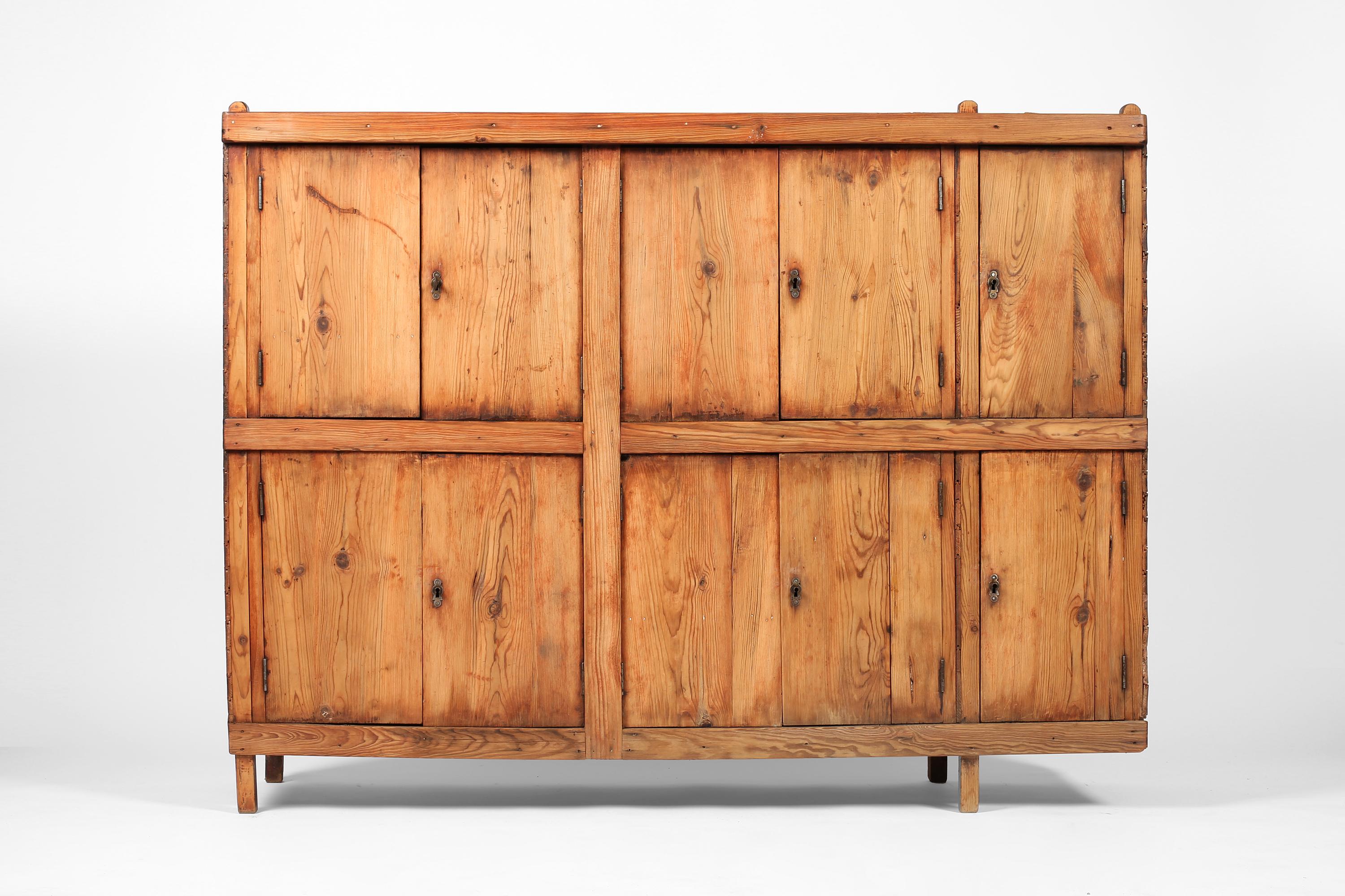 A charming early 20th century utilitarian cabinet in pine with characterful cantilevered form and decorative brass hardware. Four sets of double doors open to reveal ample storage within, with a further two single doors to the right. Very likely a