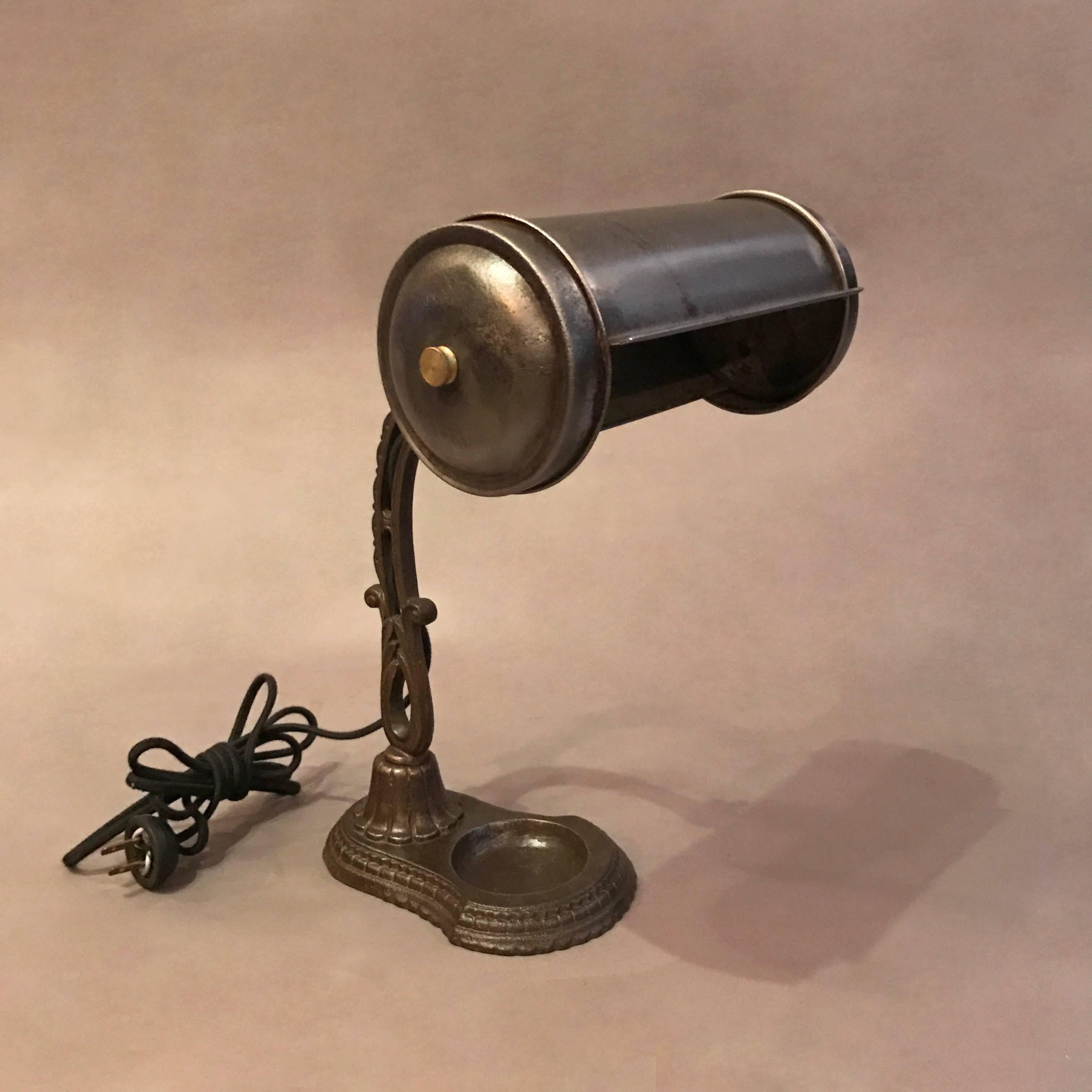 Early 20th century, library desk lamp features an ornate cast iron base and stem with a brushed steel rolled shade that articulates up and down. The lamp is newly wired with black cloth cord to accept up to a 100 watt bulb.