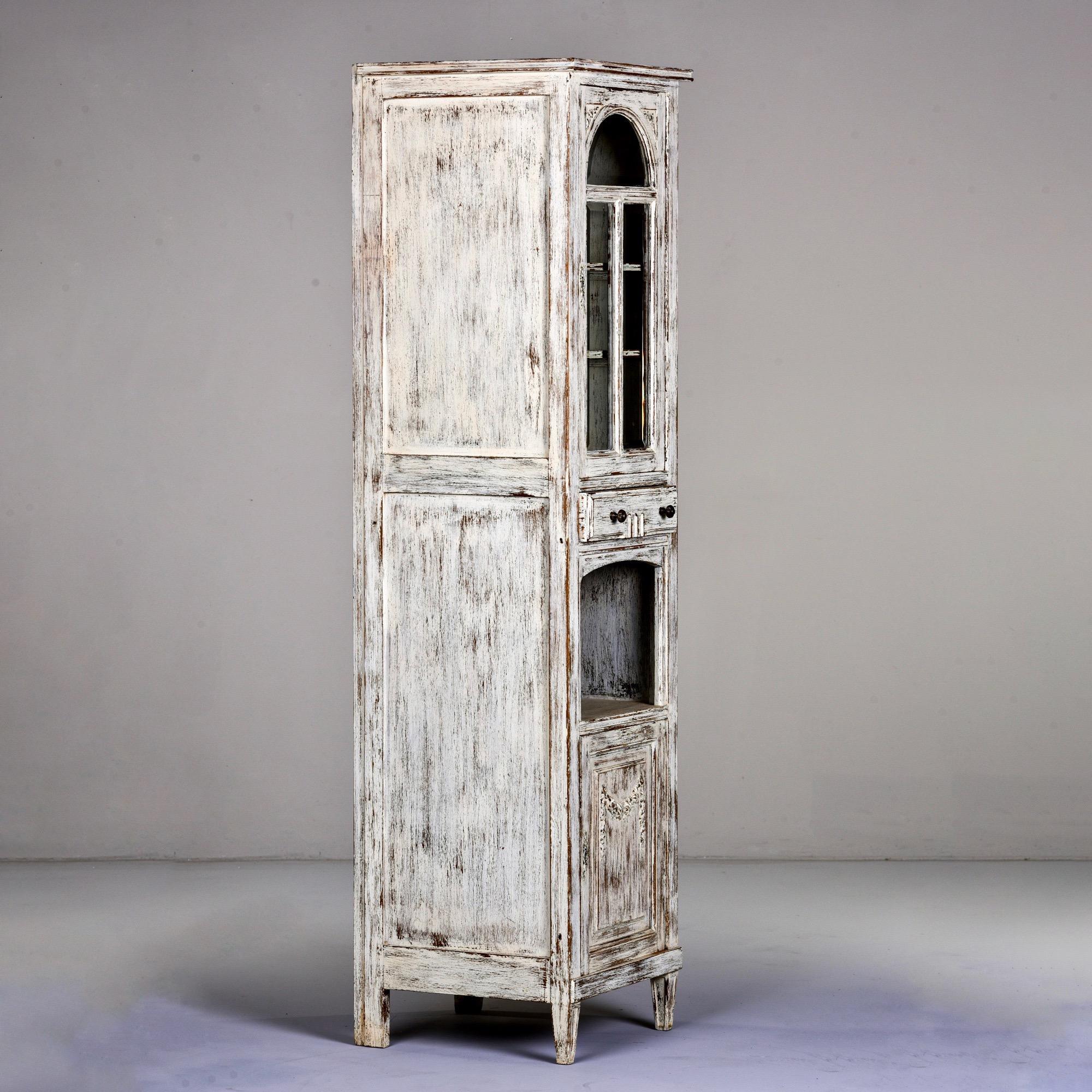 Tall and narrow French oak cabinet with white painted finish, circa 1910. Top compartment has locking glass front door and two internal shelves. In the middle is a functioning narrow drawer above an open compartment. Footed base and lower locking