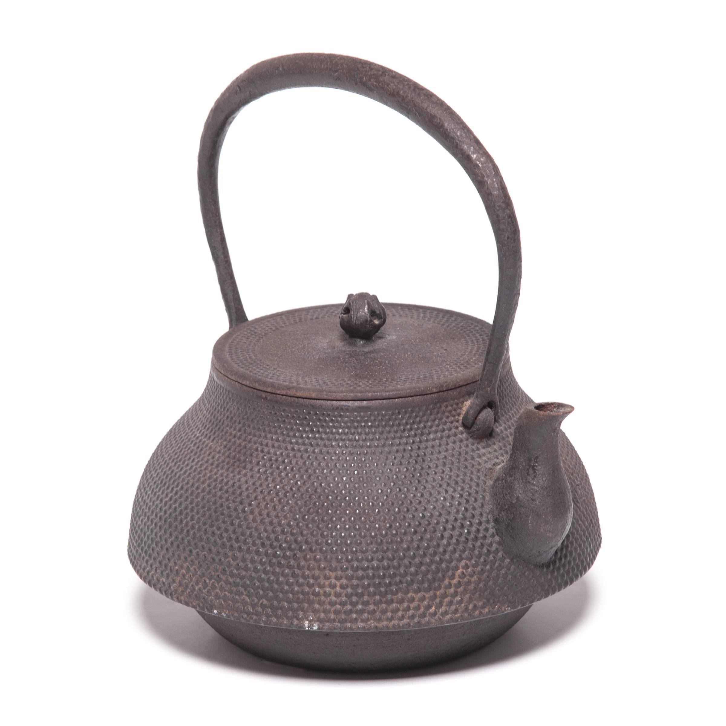 Décorée d'une surface texturée et pointillée et d'une élégante poignée arquée, cette théière japonaise était utilisée pour faire bouillir l'eau lors des cérémonies traditionnelles du thé. Connue sous le nom de tetsubin, la construction en fonte de