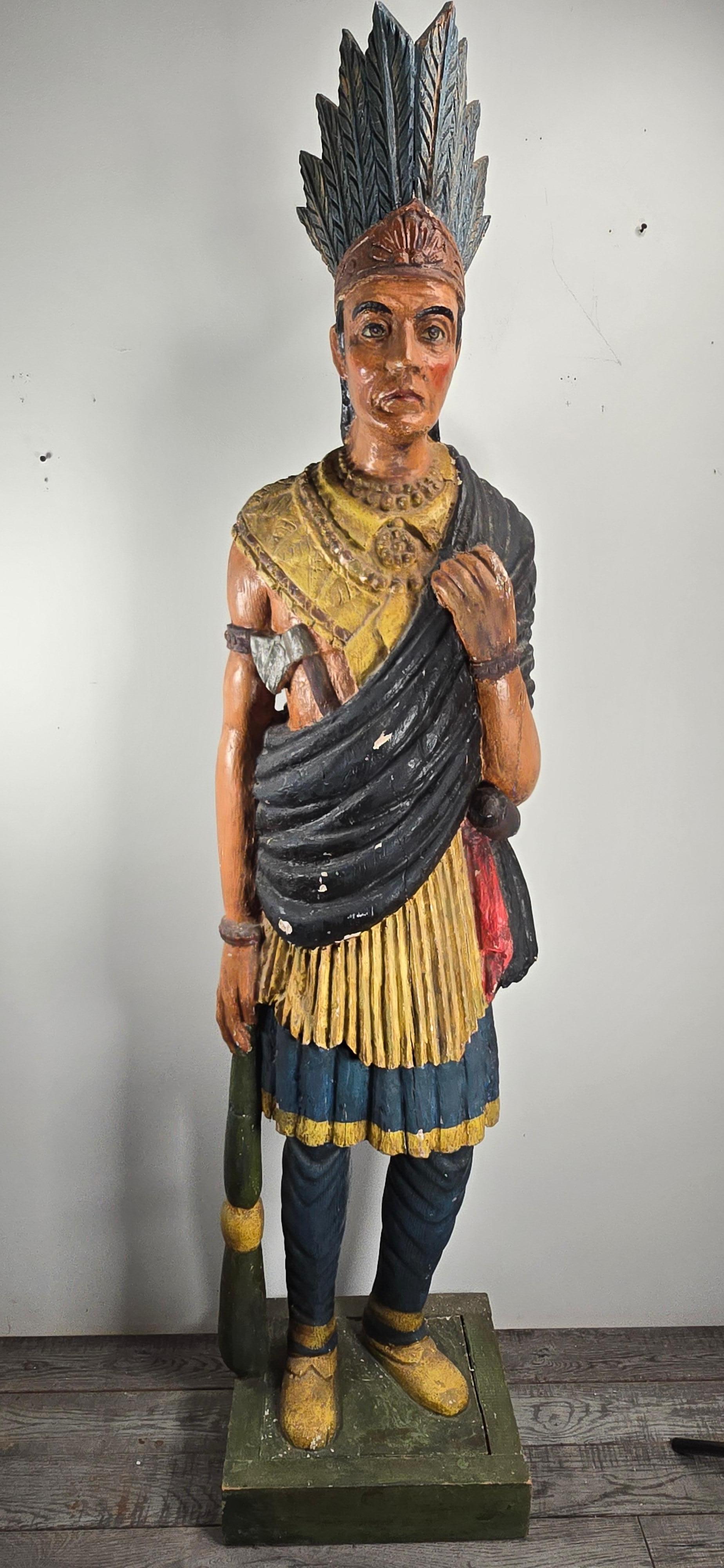 An impressive early 20th-century carved and polychrome painted cigar store Indian standing approximately 6 feet tall to the tip of the feathers. The base features replacement boards—a common and necessary repair due to the frequent rot encountered