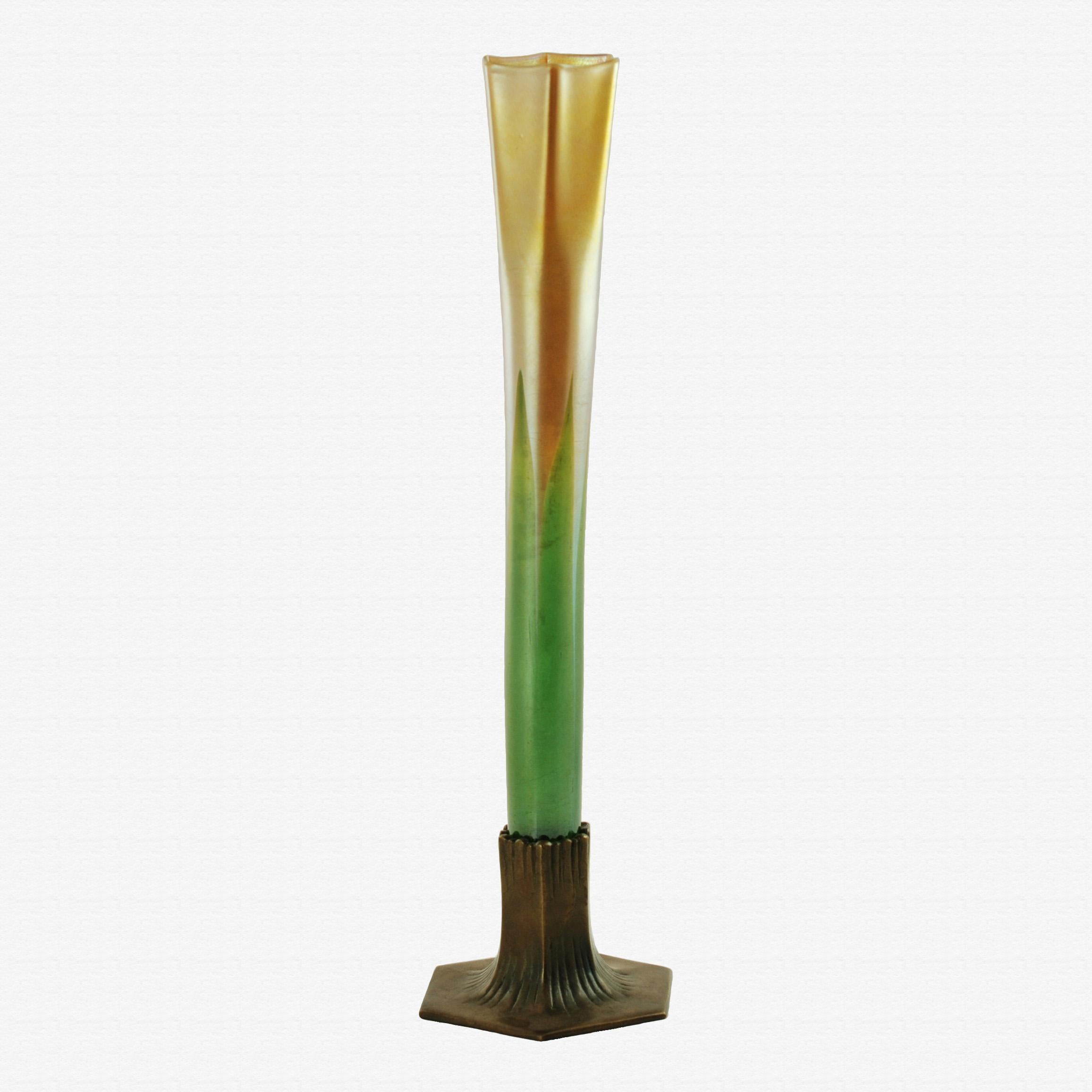 This tall Favrile glass bud vase was made by Tiffany and transitions from bottle green to a warm, iridescent gold color. The body of the vase has a cylindrical shape at the bottom which gently flares out at the top to form a ribbed mouth with six