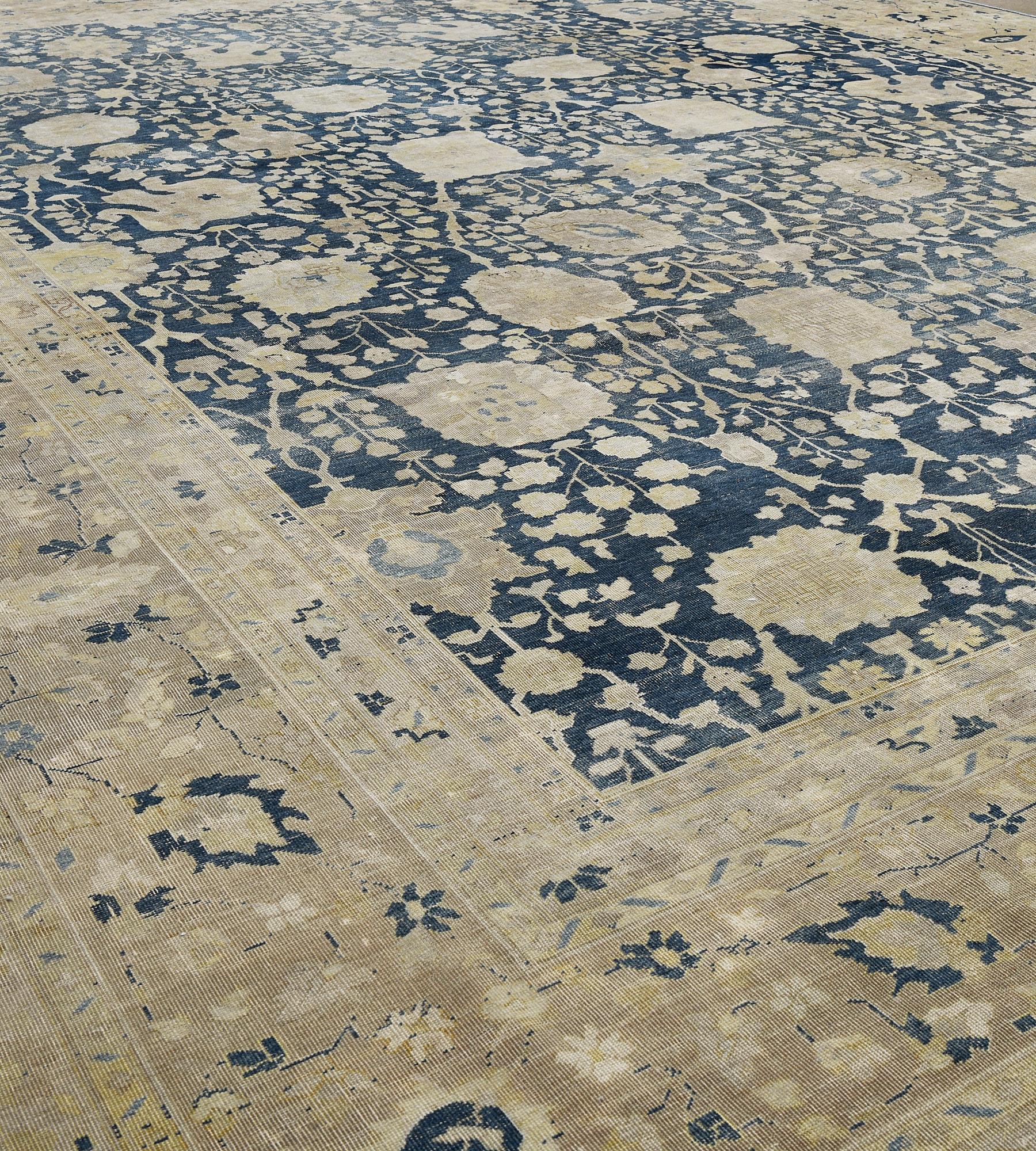 Esta alfombra tradicional persa de Tabriz, tejida a mano, tiene un campo general añil de caprichosas enredaderas de palmetas que emiten un denso motivo floral, en un borde tonal beige de enredaderas de palmetas, entre una profusión de rayas de
