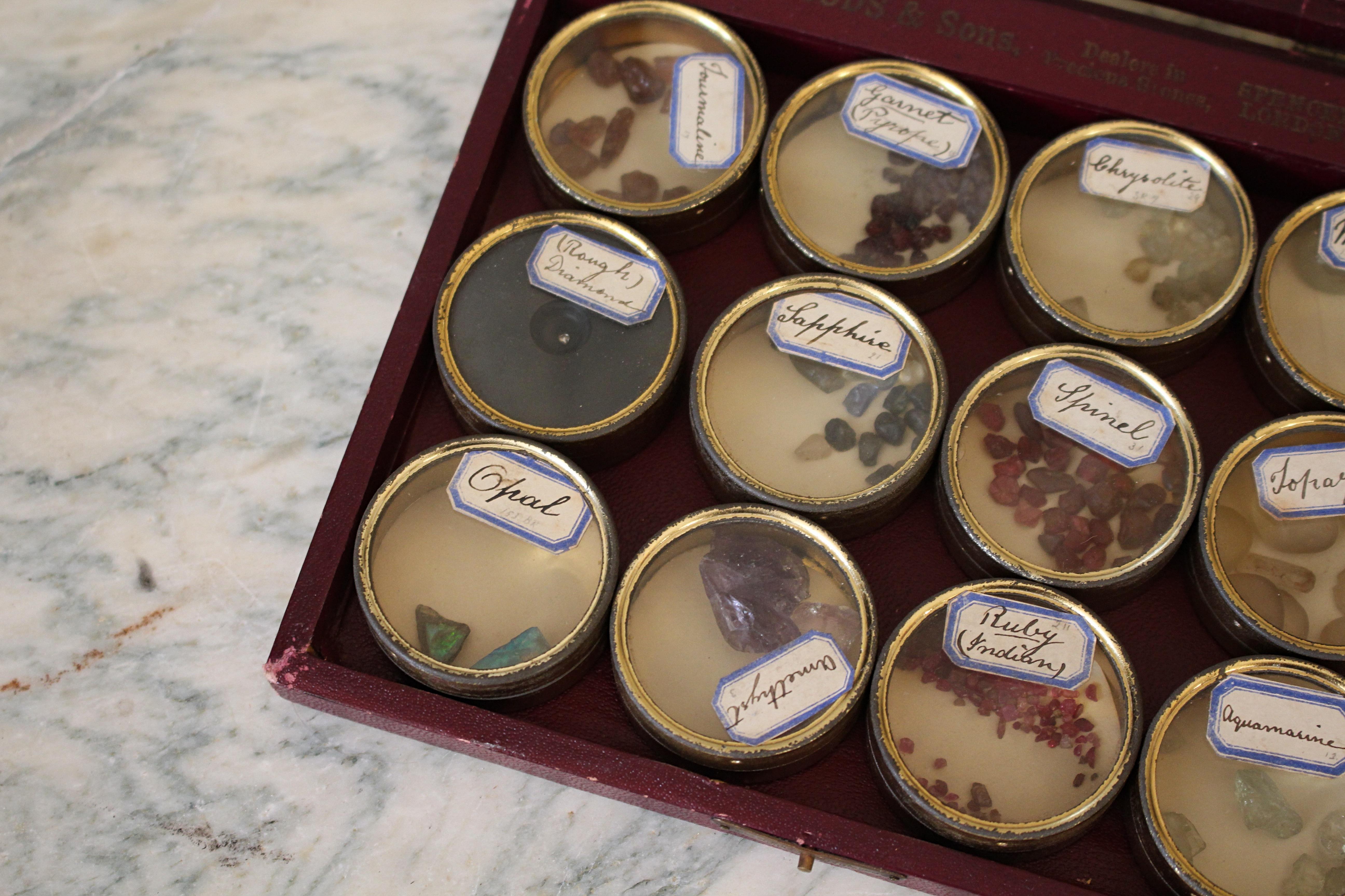 A glazed sample case of precious stones, the collection of 12 gilt tins house a range of uncut stone samples all with their original hand penned labels. Among them is a rough diamond, Indian rubys, opals, sapphires, moonstone, amethyst, garnet,