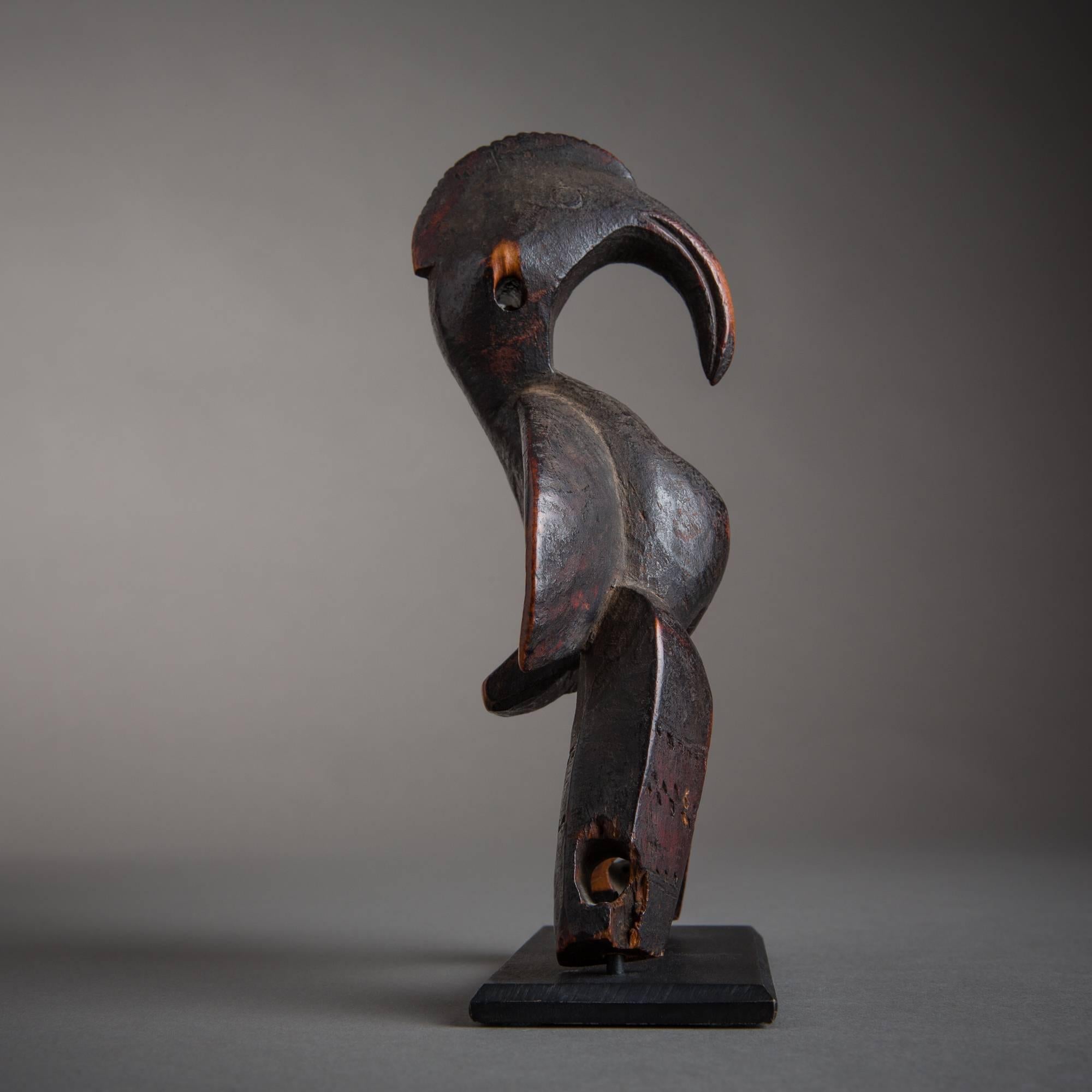 A deeply patinated pulley depicting an avian figure with anthropomorphic characteristics, dominated by an elongated, overhanging head with gracefully curved beak. The concavity of the figure’s reverse side contrasts strongly with the obverse, from