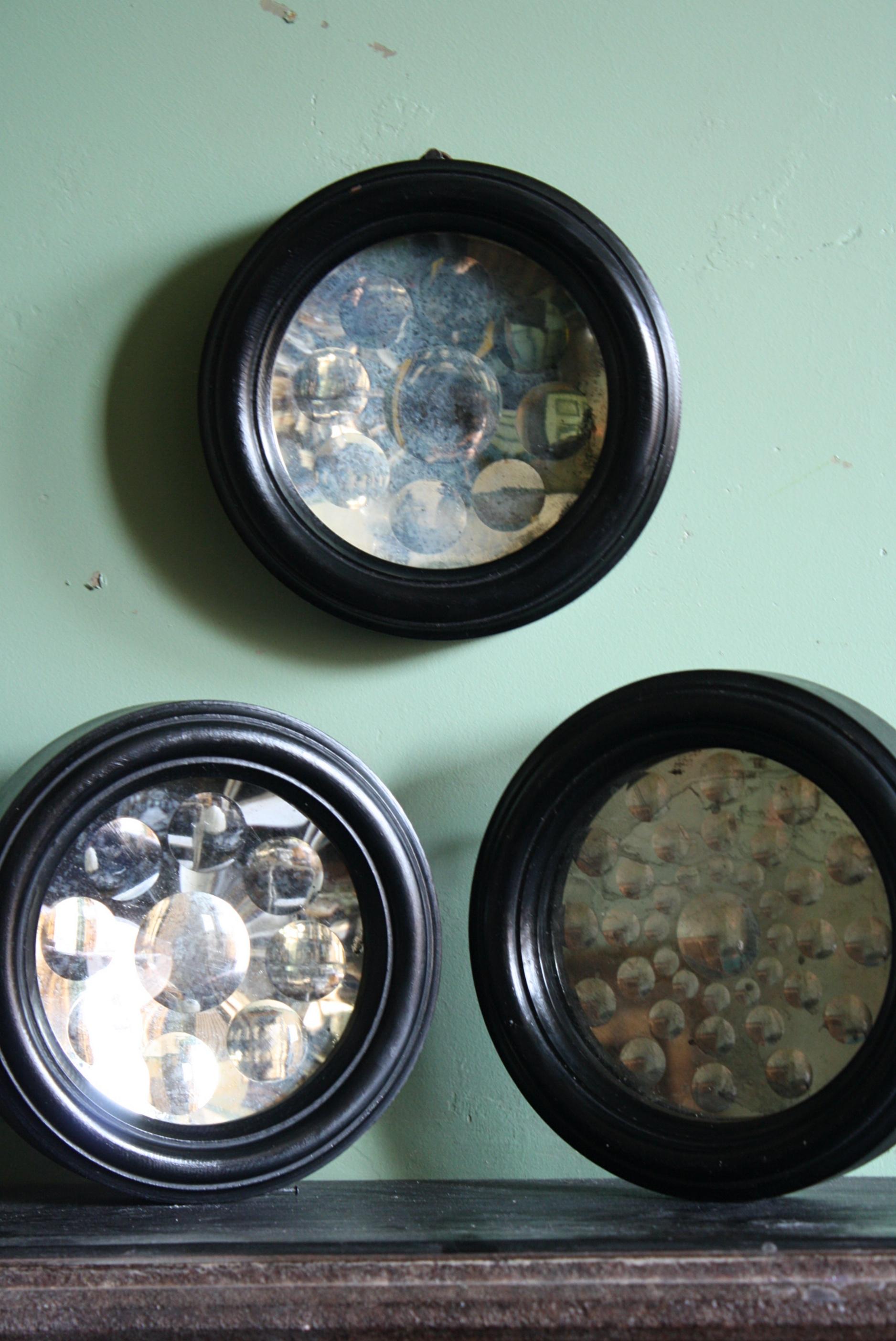 A trio of optical mirrors, housed in ebonized turned frames. Each mirror has an array of reverse ground optical lenses that throw light. The matching pair have a slight concave mirror plate with nine convex lenses the other has a more elaborate