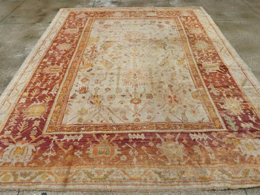 An antique Turkish Oushak room size carpet. This western Anatolian workshop carpet is patterned in a derivative Persian style, with an old ivory field decorated with ragged Harshang type palmettes and jagged leaves, and an abrashed brick main border