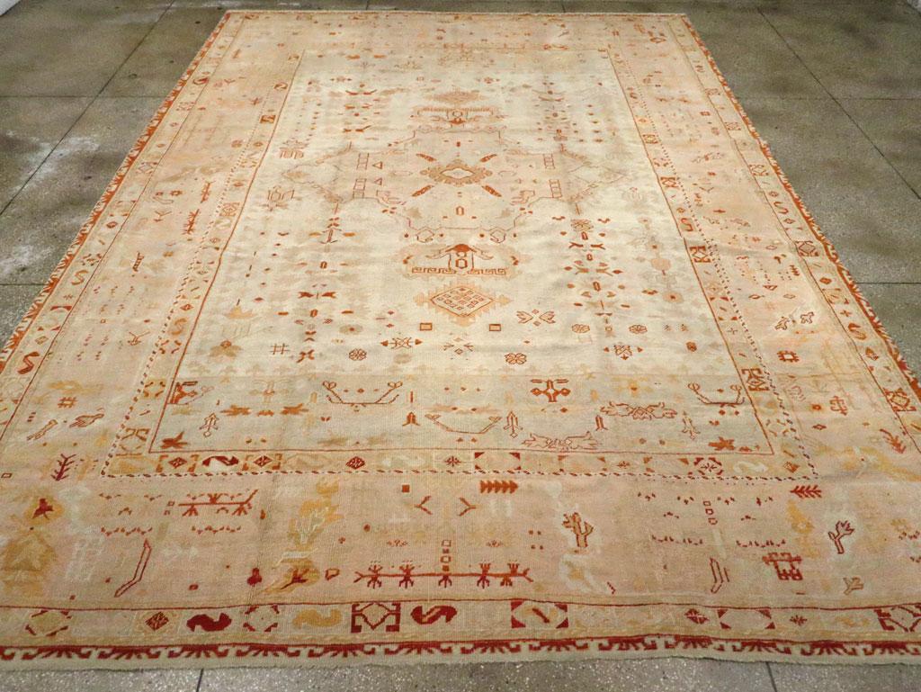 Una antigua alfombra turca Oushak tamaño habitación. El campo marfil centra un medallón beige de cuatro brazos con colgantes estilizados en medio de un relleno disperso de cadenas florales y otras formas vegetales. Un panel beige cruza cada extremo