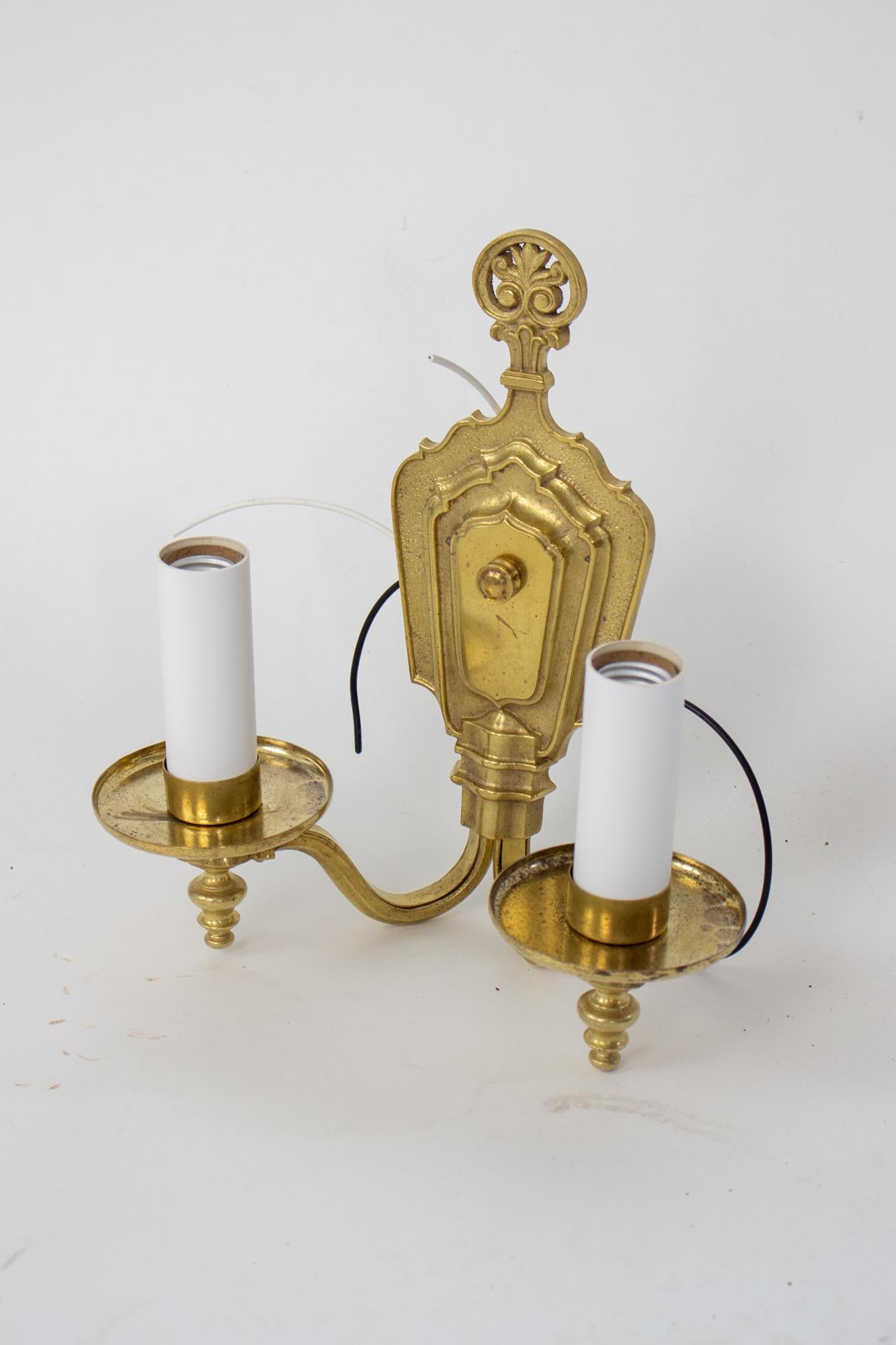 Early 20th Century two arm brass miller sconce. Cast backplate with a shield and plume pattern. Oval tubing form the arms with brass cups enclosing the sockets.