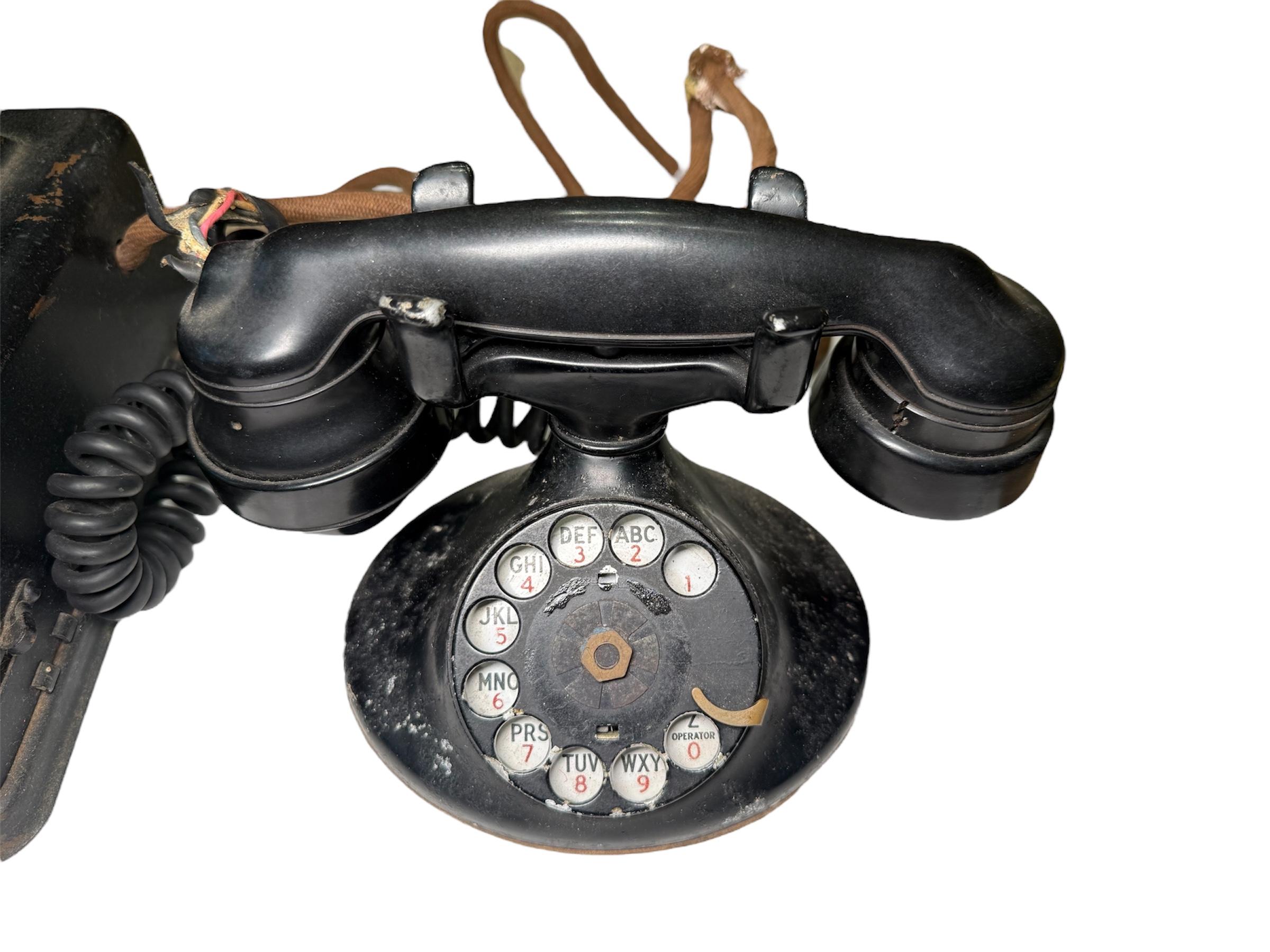Il s'agit d'un téléphone de bureau électrique WESTERN. Elle représente un téléphone de couleur noire avec une base ovale et un combiné en bakélite. La base est équipée d'un cadran rotatif. Le téléphone est relié par une Corde à une boîte métallique