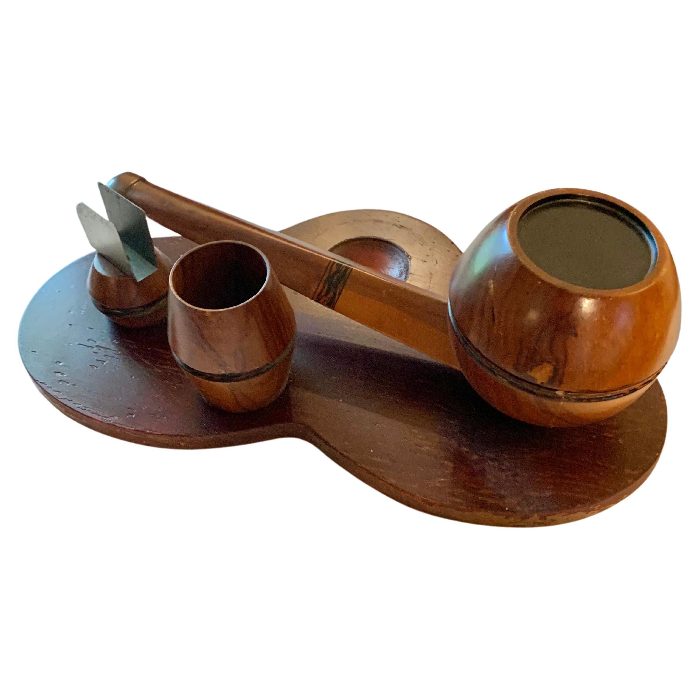 Este soporte para pipas de estilo Art Decó de principios del siglo XX se fabricó en madera a principios de la década de 1900. La pieza presenta una gran pipa con incrustaciones de diseños decorativos y una tapa de latón con bisagras que se abre para
