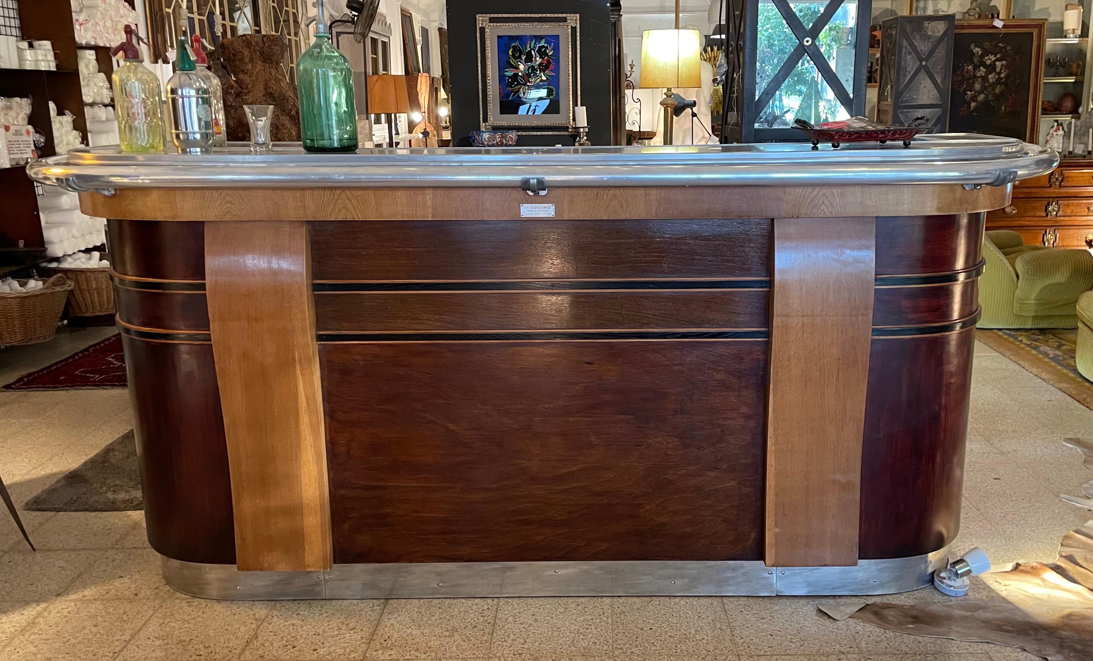 Bar counter from early 20th century. 
Rounded L-shaped, painted in dark brown but leaving its natural wood color in the two column-shaped reliefs, two marquetry inlays and under counter. 
Made of oak wood. The top surface, hand rail and baseboard