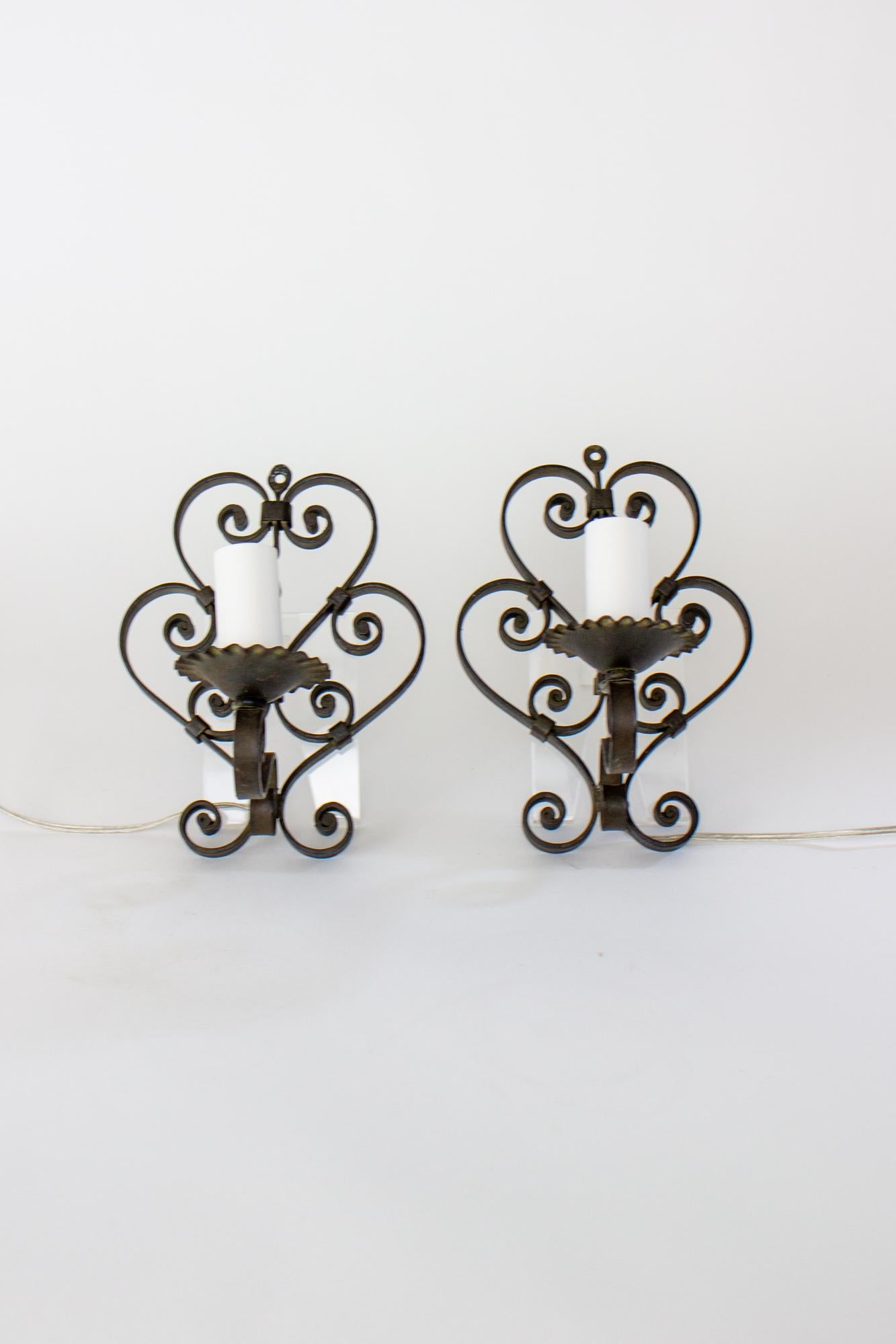 Early 20th century wrought iron pin up sconces. Wrought iron in a double heart design with curved tendrils. Original scalloped bobeche. Instead of closing the open look to enable hard wiring, these are wired to plug in, so they can be hung simply