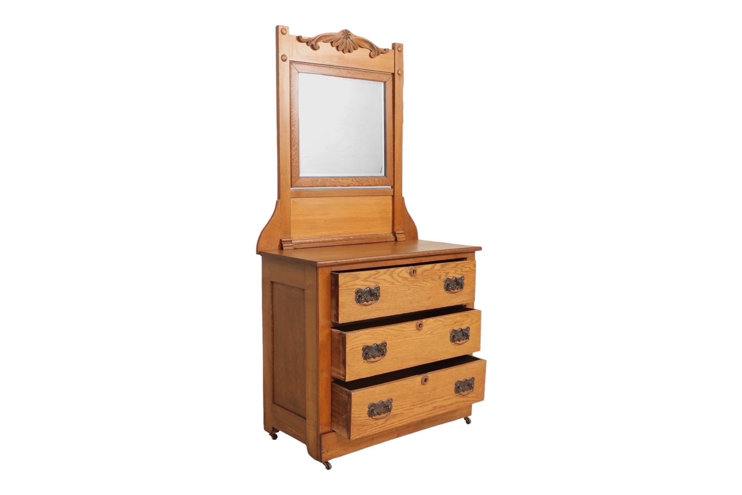 A three drawer dresser made of oak with a vanity mirror dating from the early 20th century. The square beveled mirror is adjustable, topped with a simple carved appliqué. Three slot dovetailed drawers below, open with rudimentary metal bail handles
