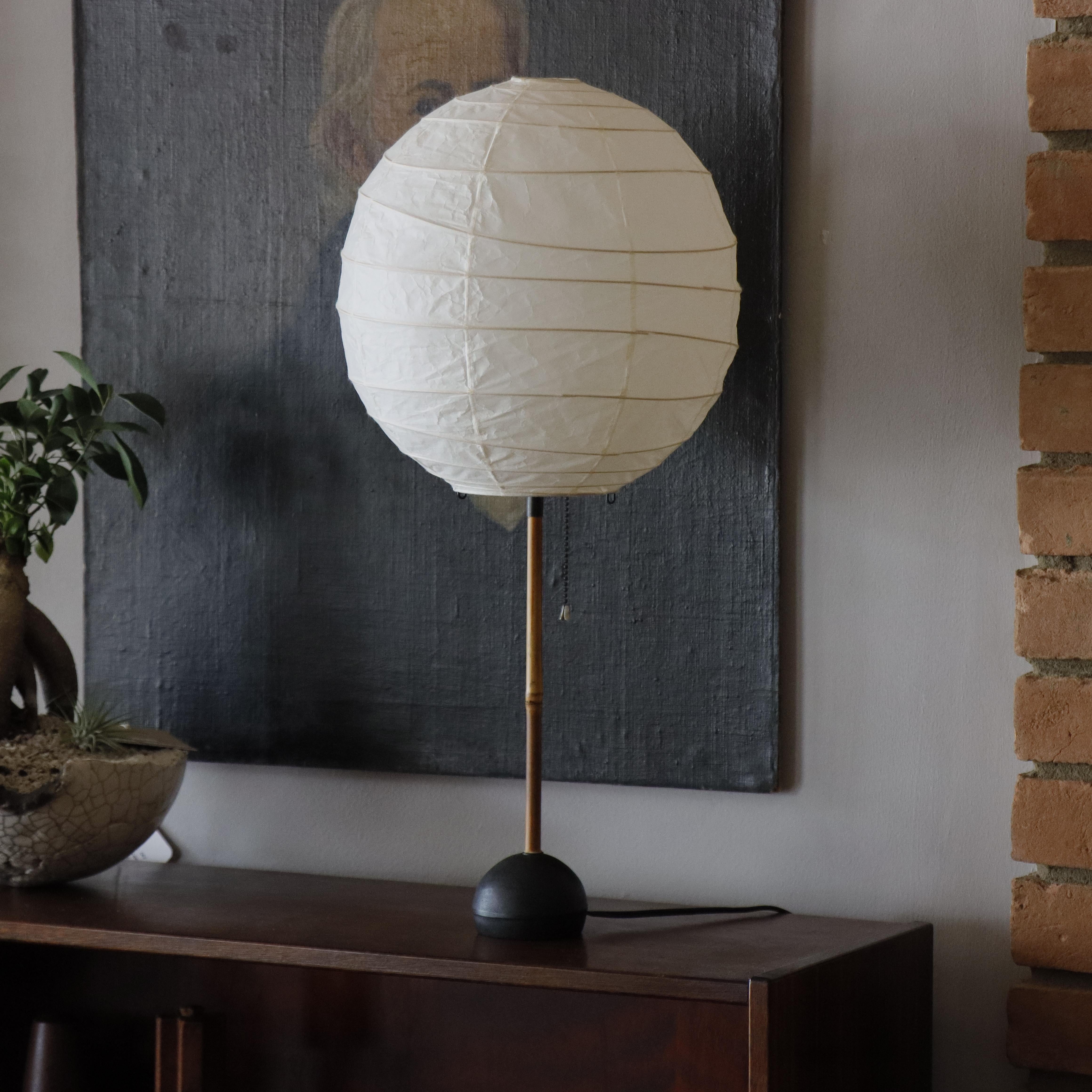 Una icónica escultura de luz Akari diseñada por Isamu Noguchi, maestro de la forma y la luz. Esta pieza vintage conserva su cordón y herrajes originales, ofreciendo autenticidad y elegancia atemporal. Hecha a mano con papel washi y cintas de bambú,
