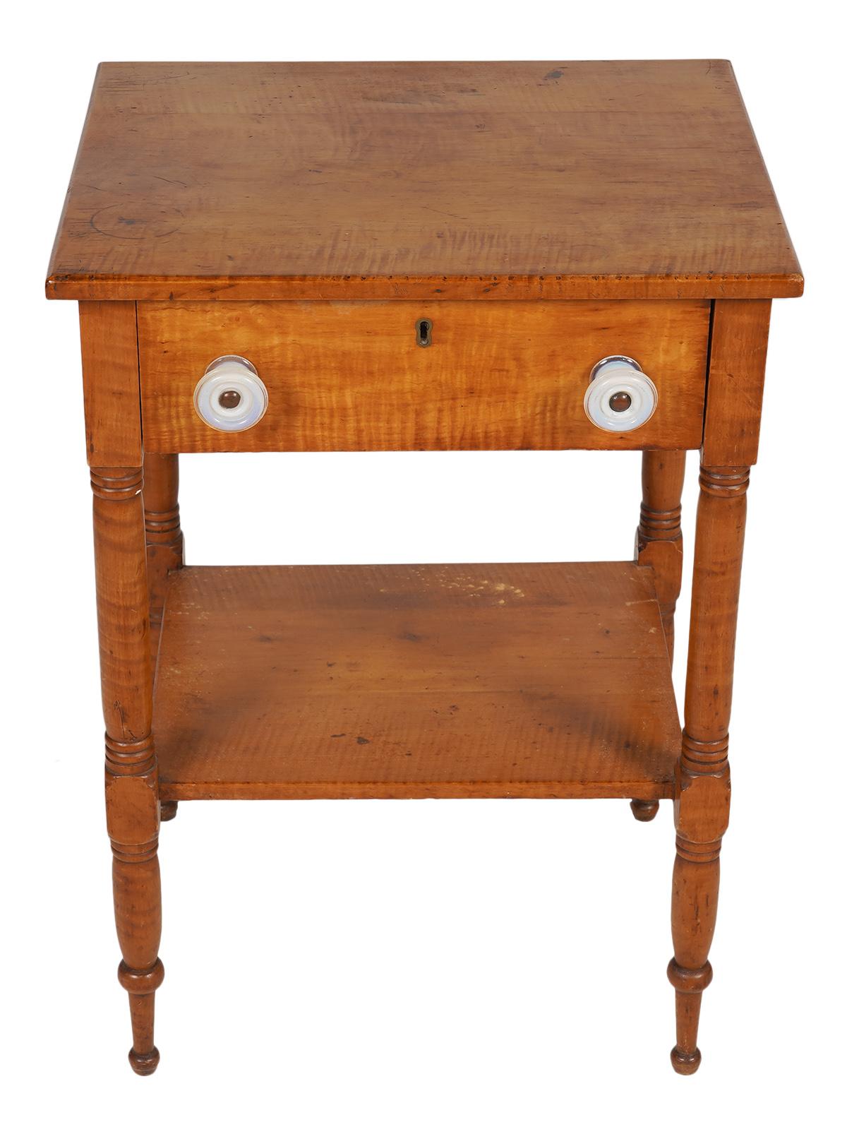 This early American tiger maple work table or stand features a top with slightly rounded edges above a frieze holding a drawer with likely original milk glass pulls. The four turned and carved legs are united by a lower shelf.