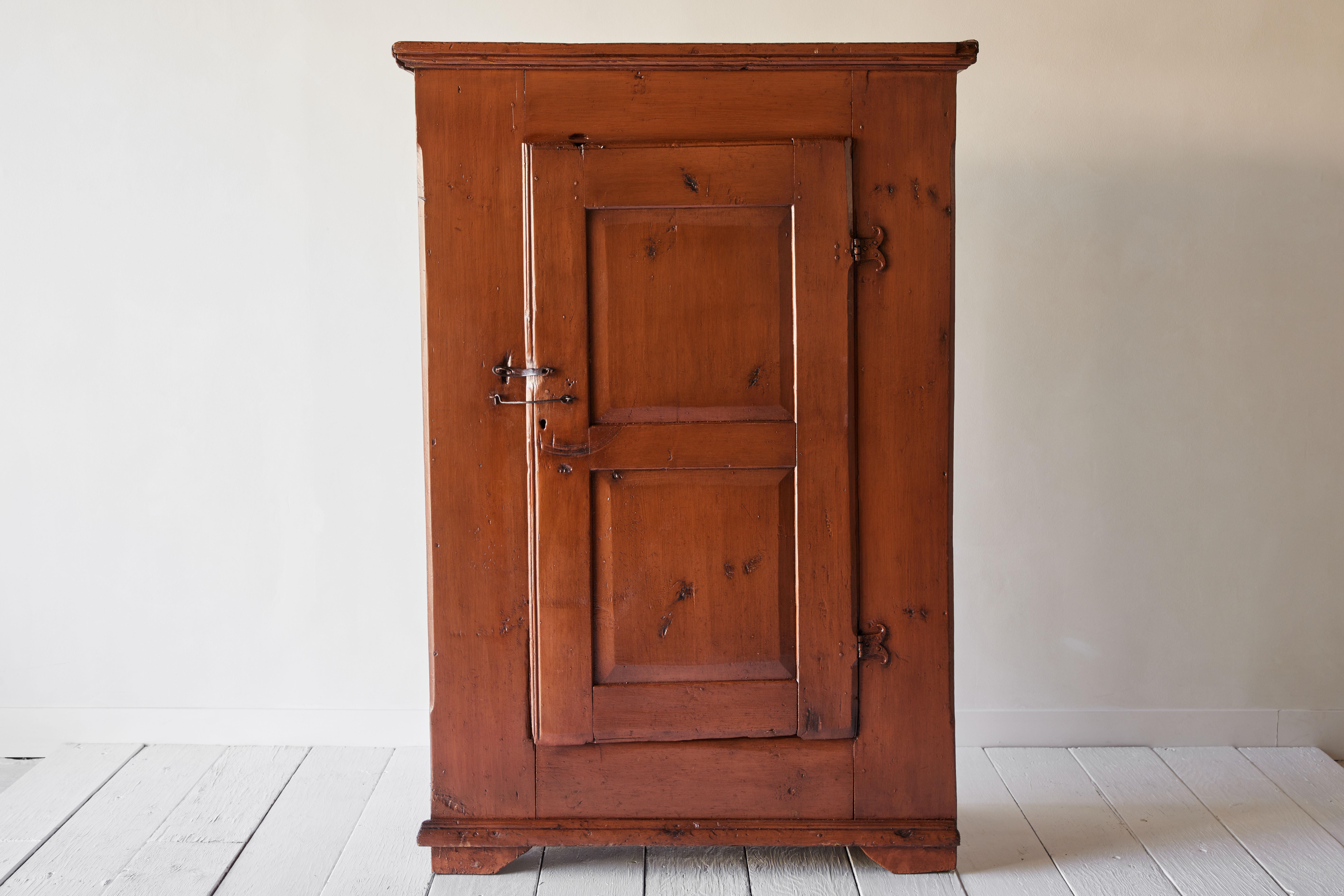 A very handsome rustic early American single door cupboard painted rusty orange with dark colored undertones. This cabinet is a wonderful utilitarian piece and features an open interior allowing room for ample storage. The foundation is there if you