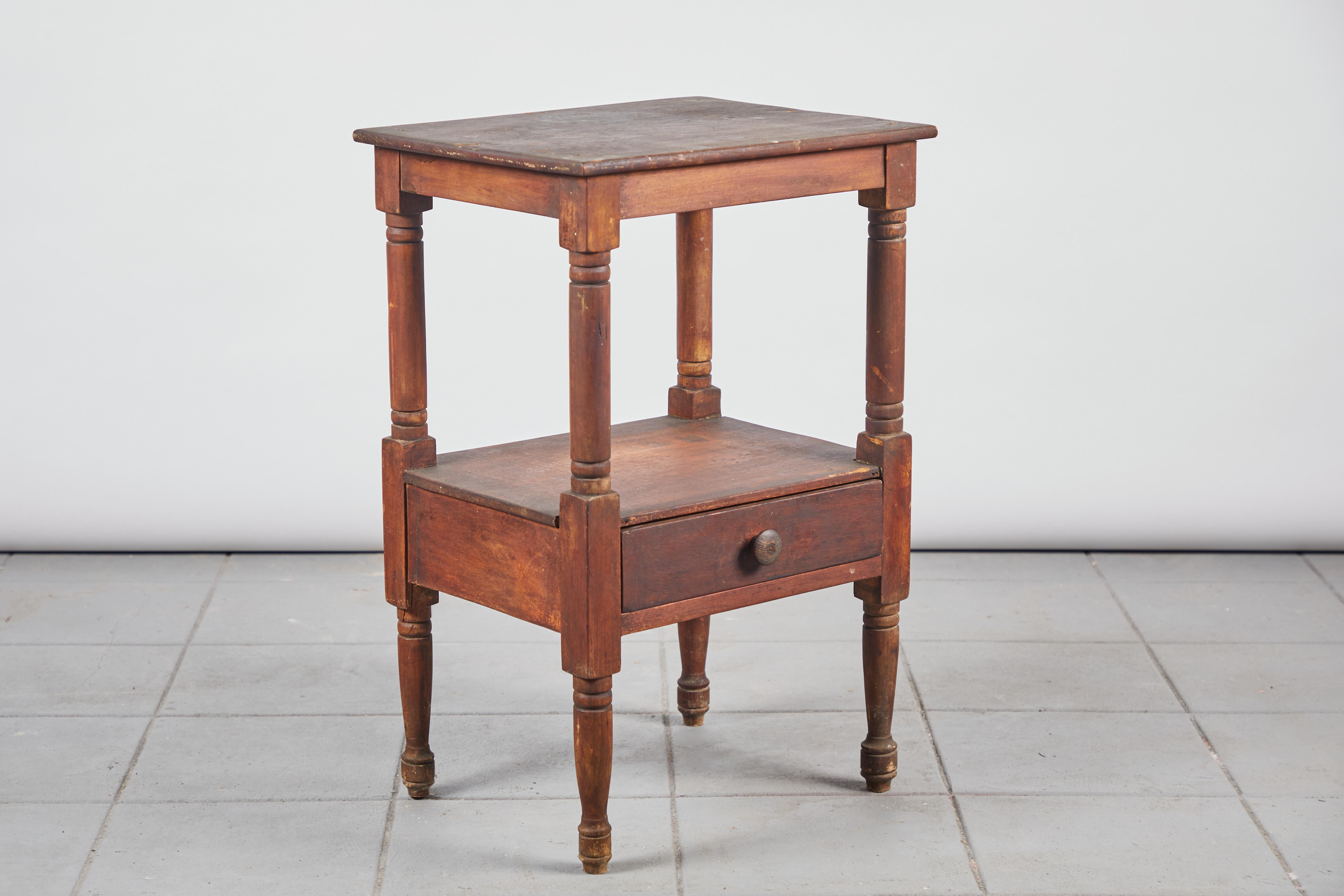 Early American rustic side table with turned legs and a lower shelf that offers a single drawer.
