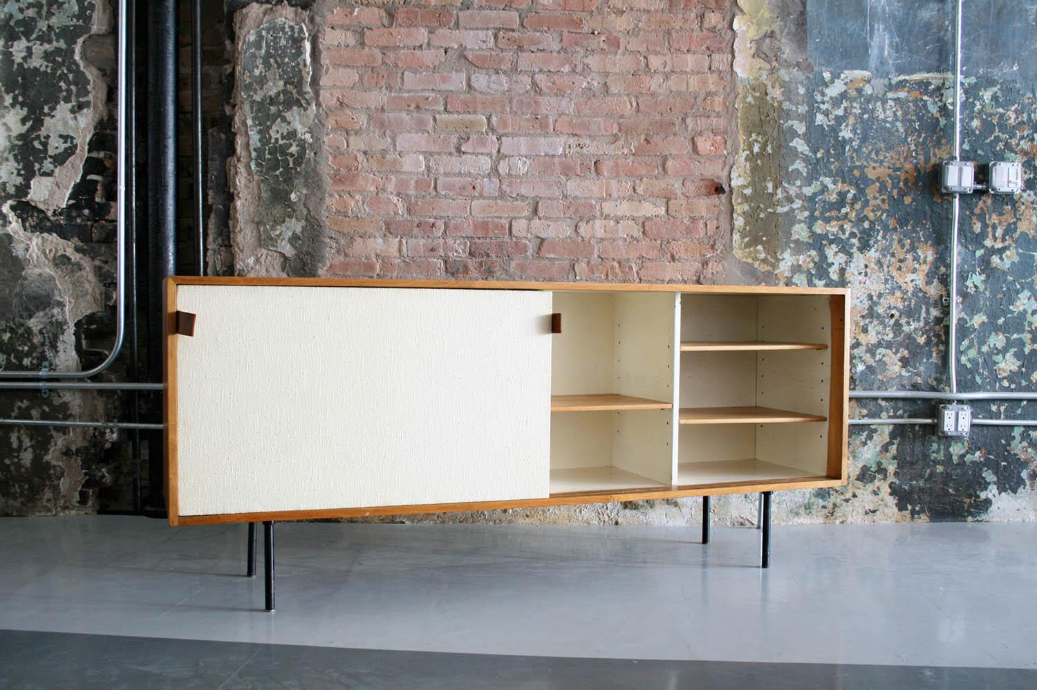 This credenza designed by Florence Knoll, features handsome original fabric covered sliding doors with leather pulls. Inside are five shelves and one sliding drawer. The bleached walnut case sits on a steel base. No maker's mark is visible on this