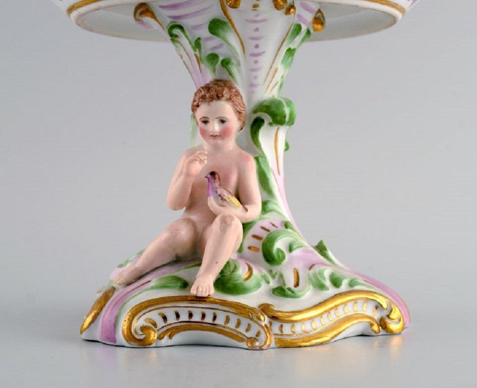 Early and rare Royal Copenhagen compote in openwork porcelain with hand-painted flowers and gold decoration. 
Foot modelled with Putto holding a bird. 
Museum quality. 
Early 19th century.
Measures: 22 x 19 cm.
In excellent condition.
Signed.