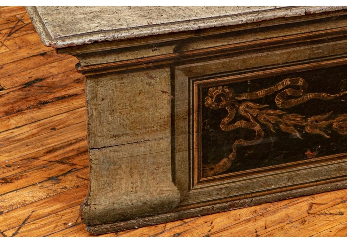 A Neoclassical decorated carved and painted bench with lift up seat for storage. The back with scrolled crest and painted with a trompe l'loeil architectural frieze with masks, ribbons and an center crest with a bird on three hills. The lower frame