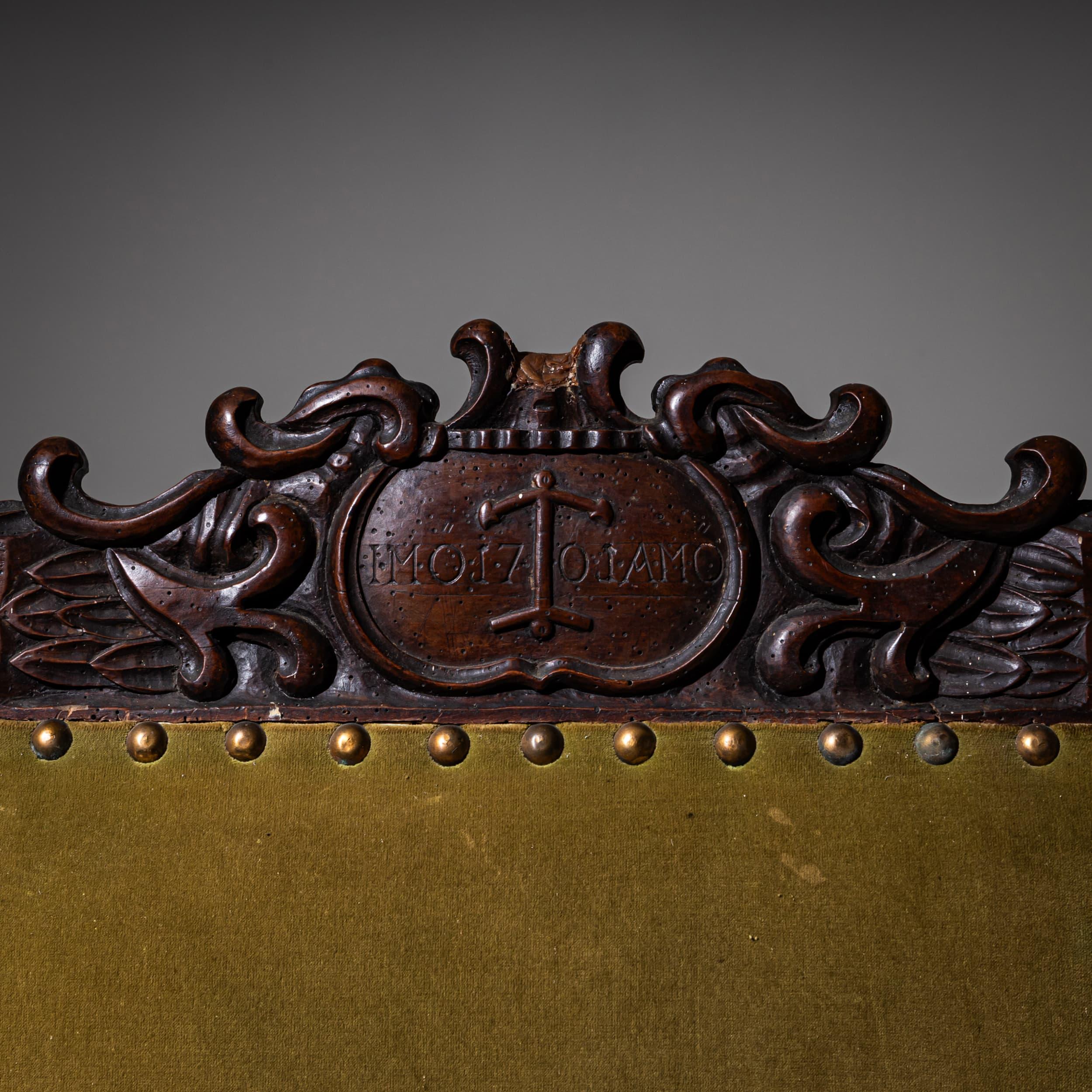 Impresionante sillón de principios del Barroco con reposabrazos rectos tapizados, patas de balaustre torneadas y crucetas. El respaldo rectangular está rematado con una crestería tallada que presenta una cartela con un motivo de ancla. La