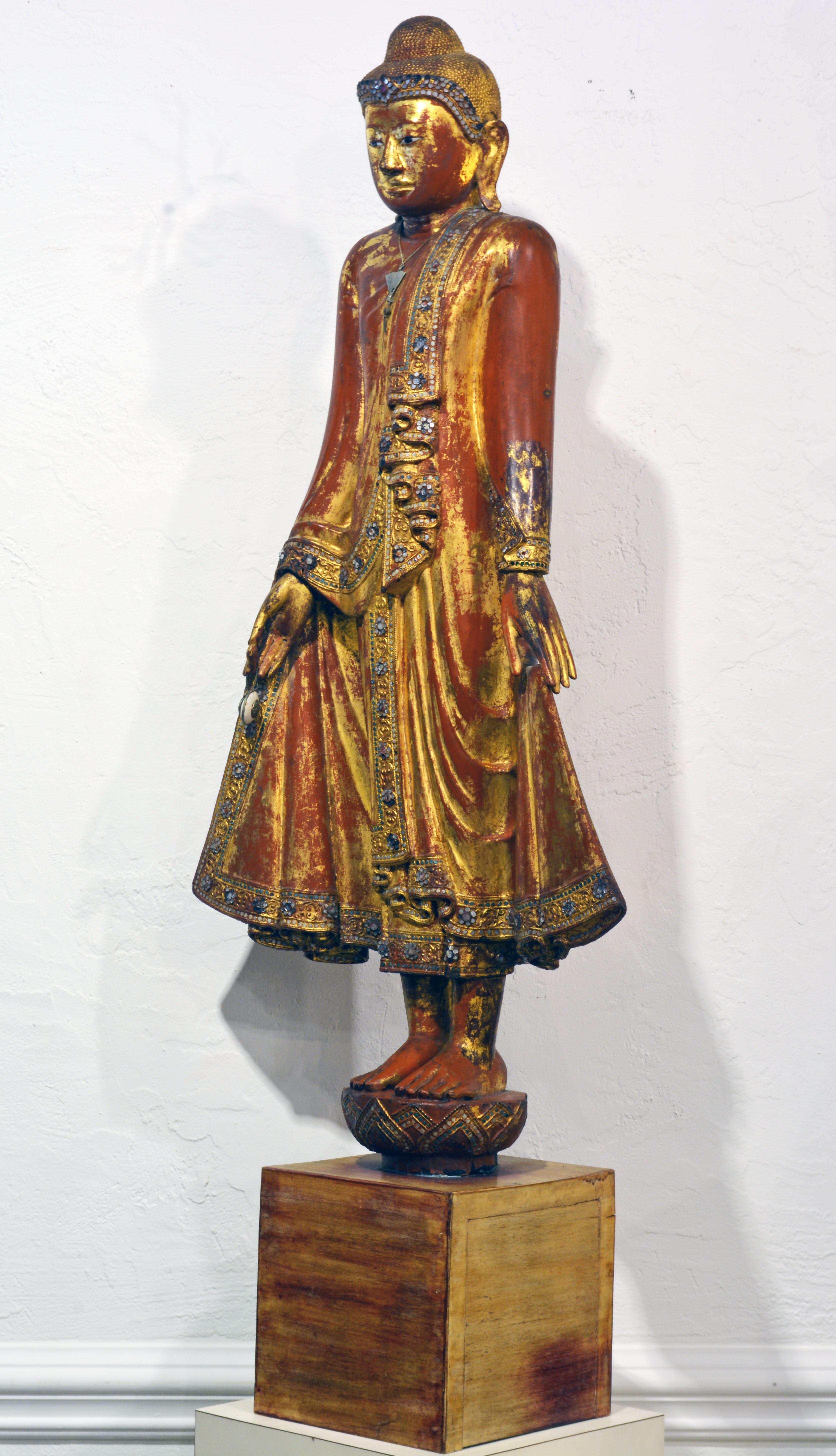 A superior 50 inches tall carved lacquer and gilt statue of the Mandalay style Shakyamuni Buddha (Myanmar) standing on a stylized lotus flower and wearing princely bejeweled robes. The beautifully carved face with porcelain or glass eyes. Standing