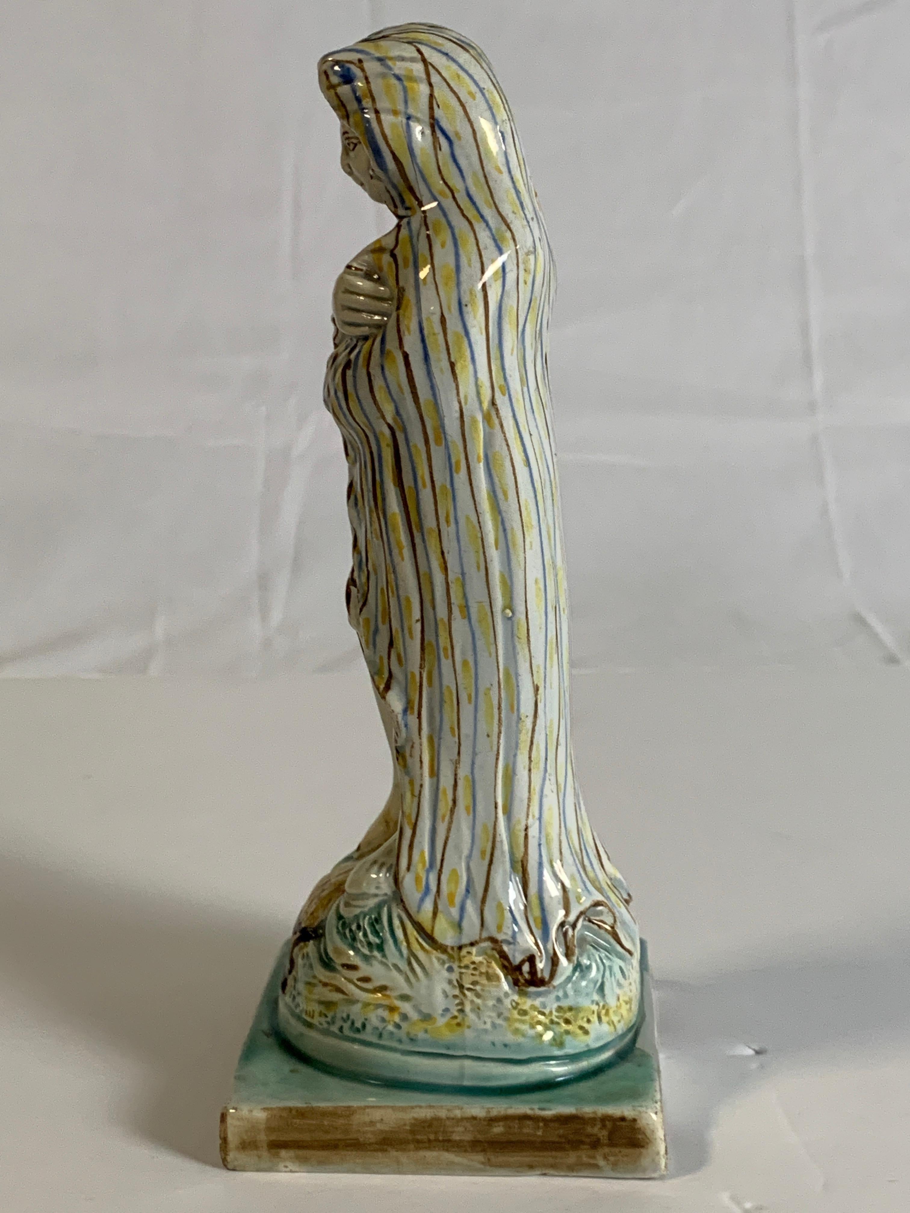 We are pleased to offer this naive and charming 18th century creamware figure of a woman personifying winter. Modeled standing on a plinth she wraps herself in a hooded cloak hand-painted with light brown and light blue stripes and spots of yellow.