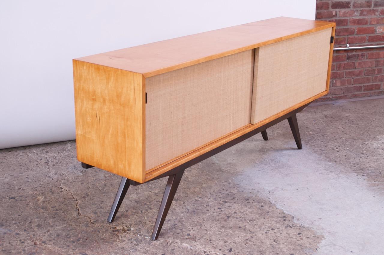 Uncommon and early Florence Knoll Credenza (1947) featuring a birch cabinet, stained mahogany base, and grasscloth-wrapped doors, which slide open to reveal three wooden shelves to the left and two plate glass shelves to the right. Dramatic lines