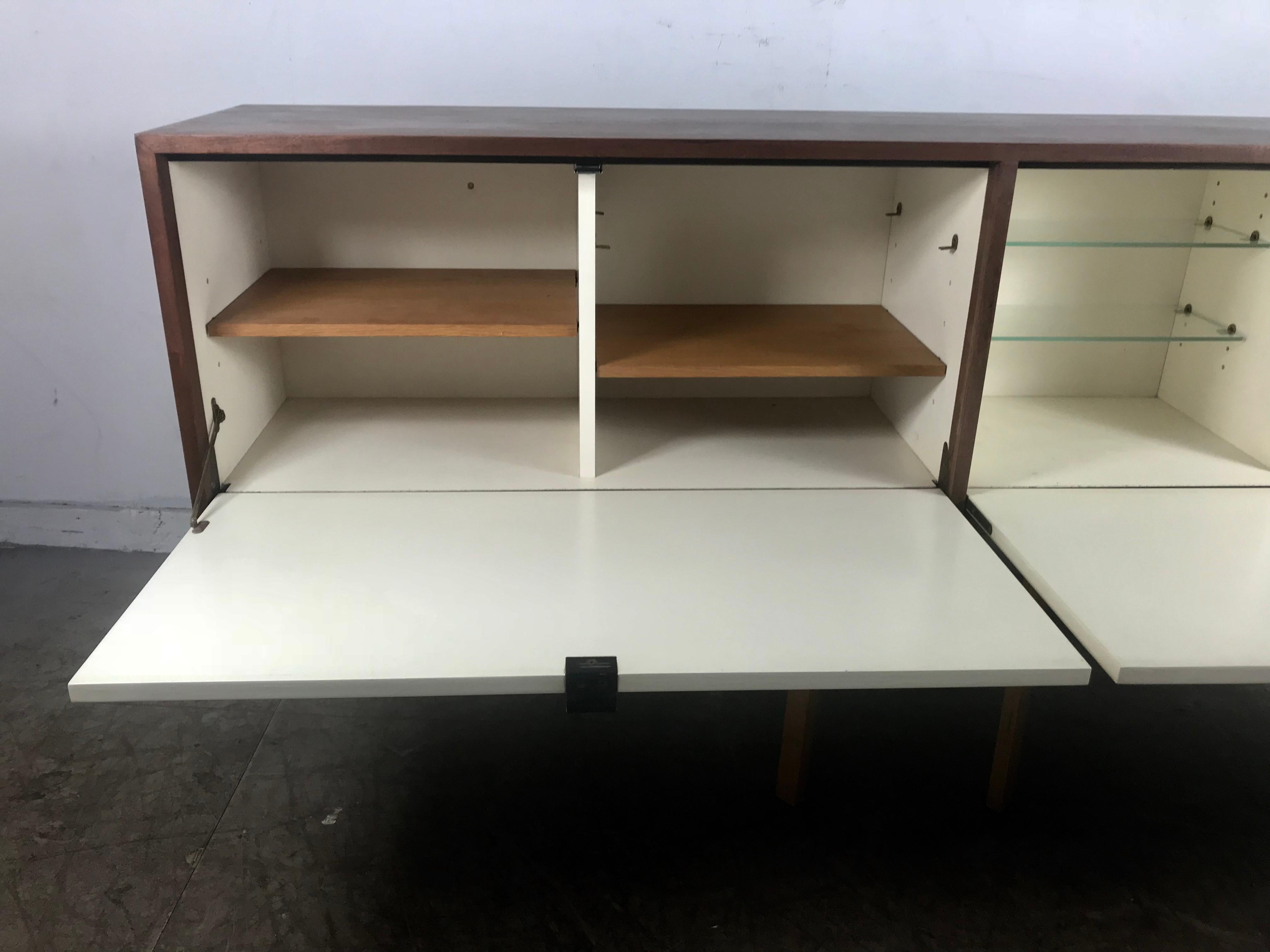 Early Florence Knoll for Knoll NY wall hung credenza or bar, walnut finish, white lacquer drop down doors, original leather pulls, featuring two oak shelves and two glass shelves. Hand delivery avail to New York City or anywhere en route from