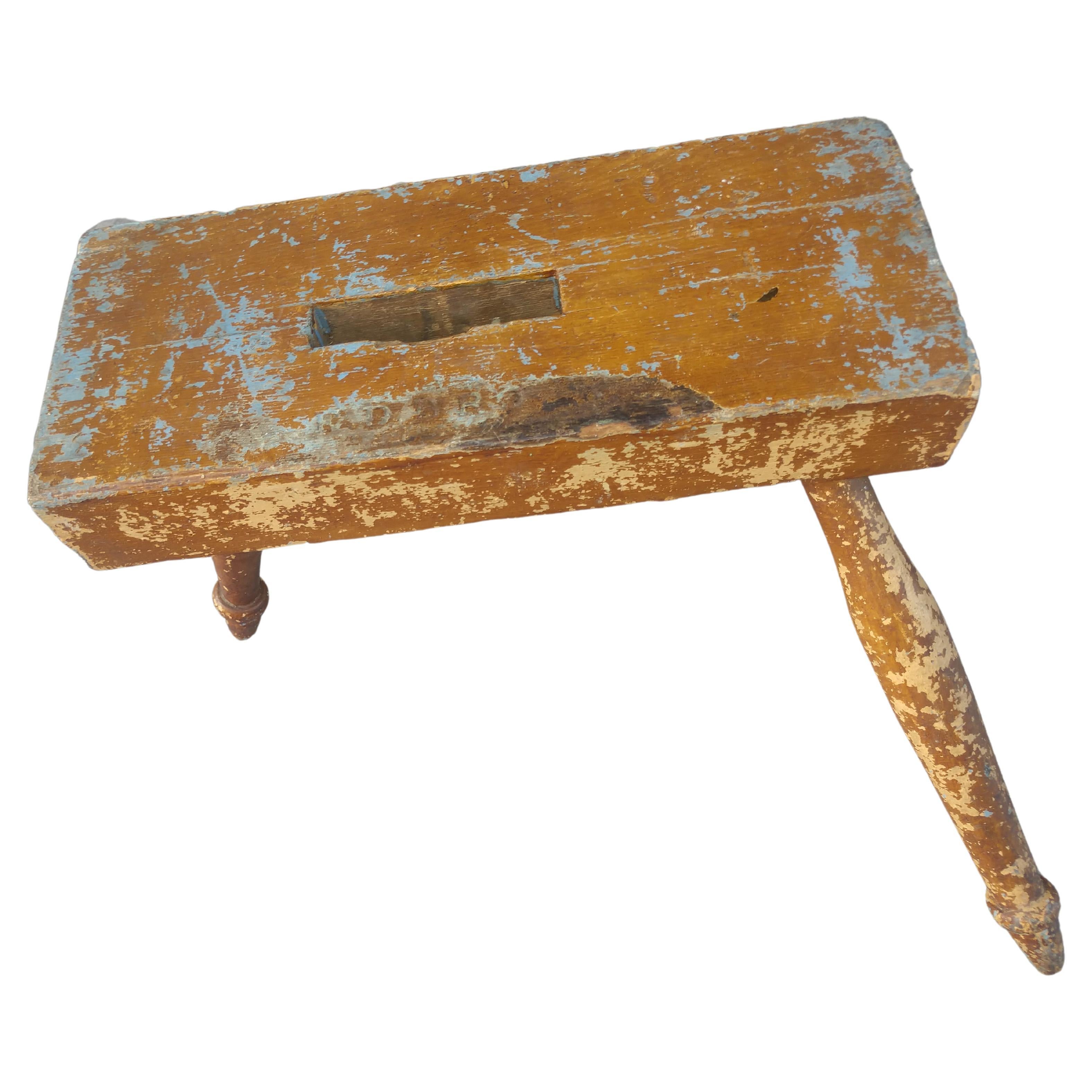 Taburete de ordeño escultórico de 3 patas con múltiples capas de pintura. Encimera rectangular con un agujero de mano cortado por el centro y apoyada en 3 patas torneadas. Hay una marca con letras en la parte superior, pero no se puede descifrar. En