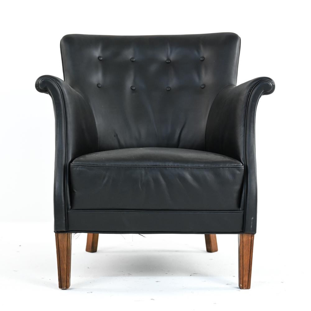 This perfectly proportioned lounge chair, designed and manufactured by Frits Henningsen, features a classic Danish tufted backrest with subtle wingtips, unusual scrolled and splayed arms, and channeled front legs. The fine quality black leather