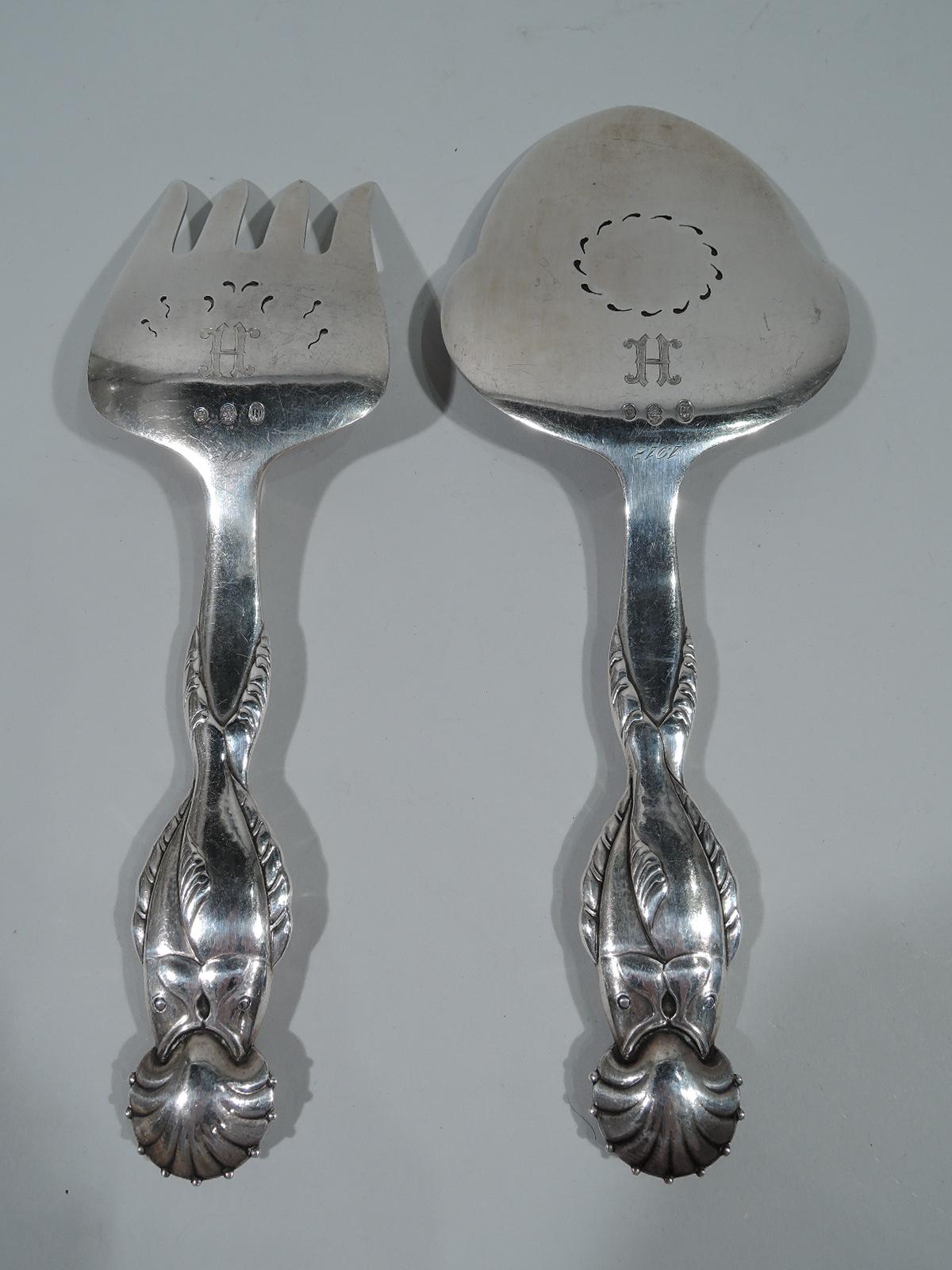 Early 830 fish serving slice and fork. Made by Georg Jensen in Copenhagen in 1925.

Each: Double-sided figural handle with entwined fish merging to form single split tail, and open mouths swallowing scallop shell terminal. Fork has curved shank