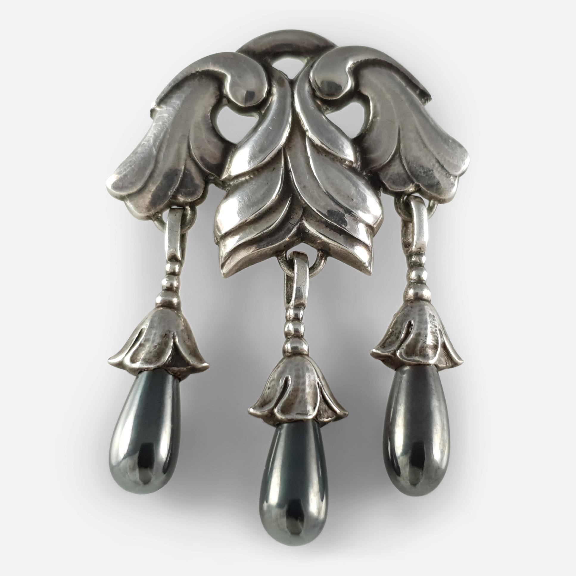 Une broche Georg Jensen en argent et hématite #132, vers 1915-1930.  La partie supérieure argentée et feuilletée suspend trois gouttes d'hématite polie. 

La broche est estampillée de la marque du fabricant GI à l'intérieur d'un cercle de perles
