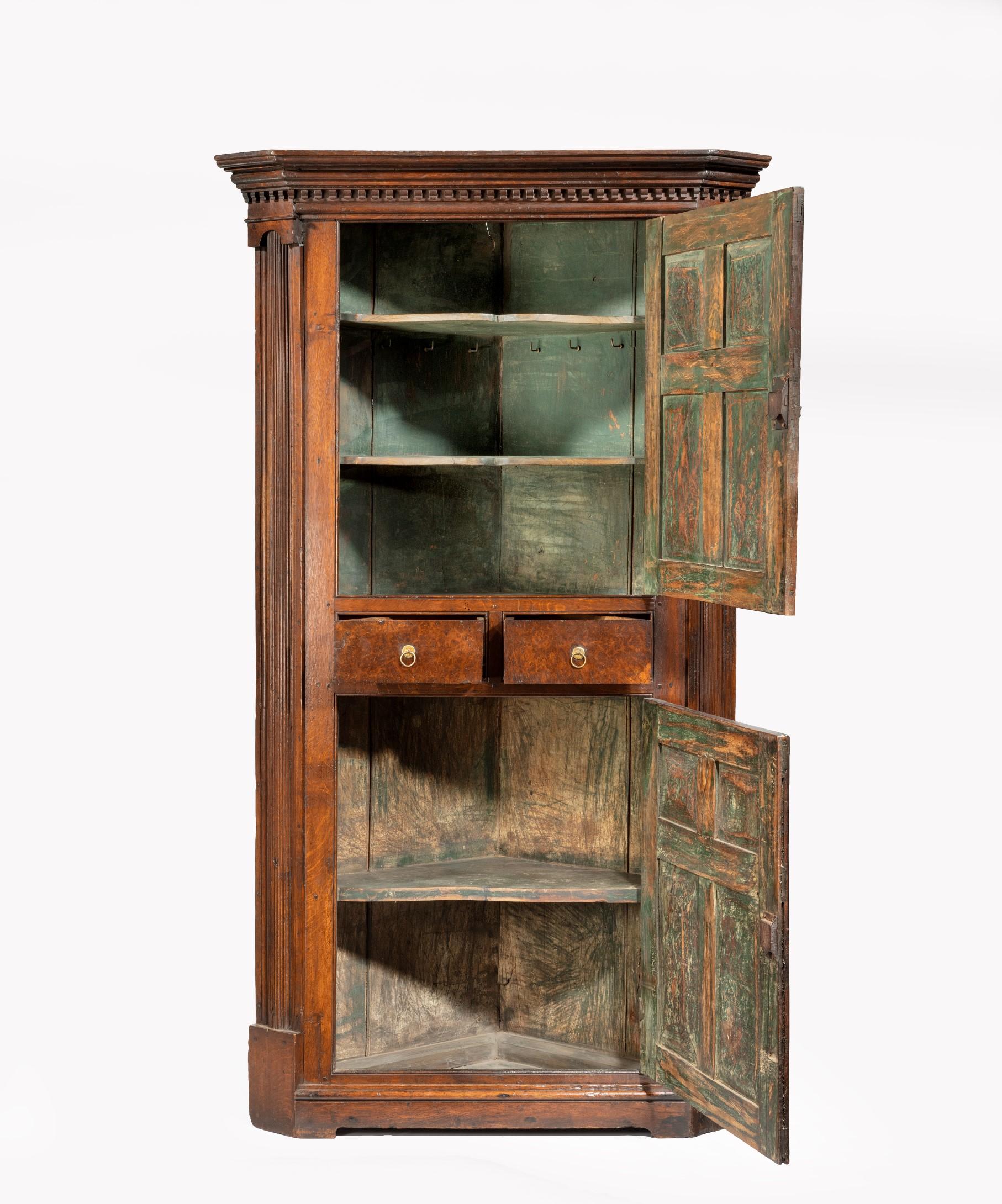 A wonderful George I oak and burr yew standing corner cupboard, the corner cupboard's dentil moulded cornice supported by a pair of fluted pilasters which flank two panelled cupboard doors, the two cupboard doors have panels of solid burr yew and